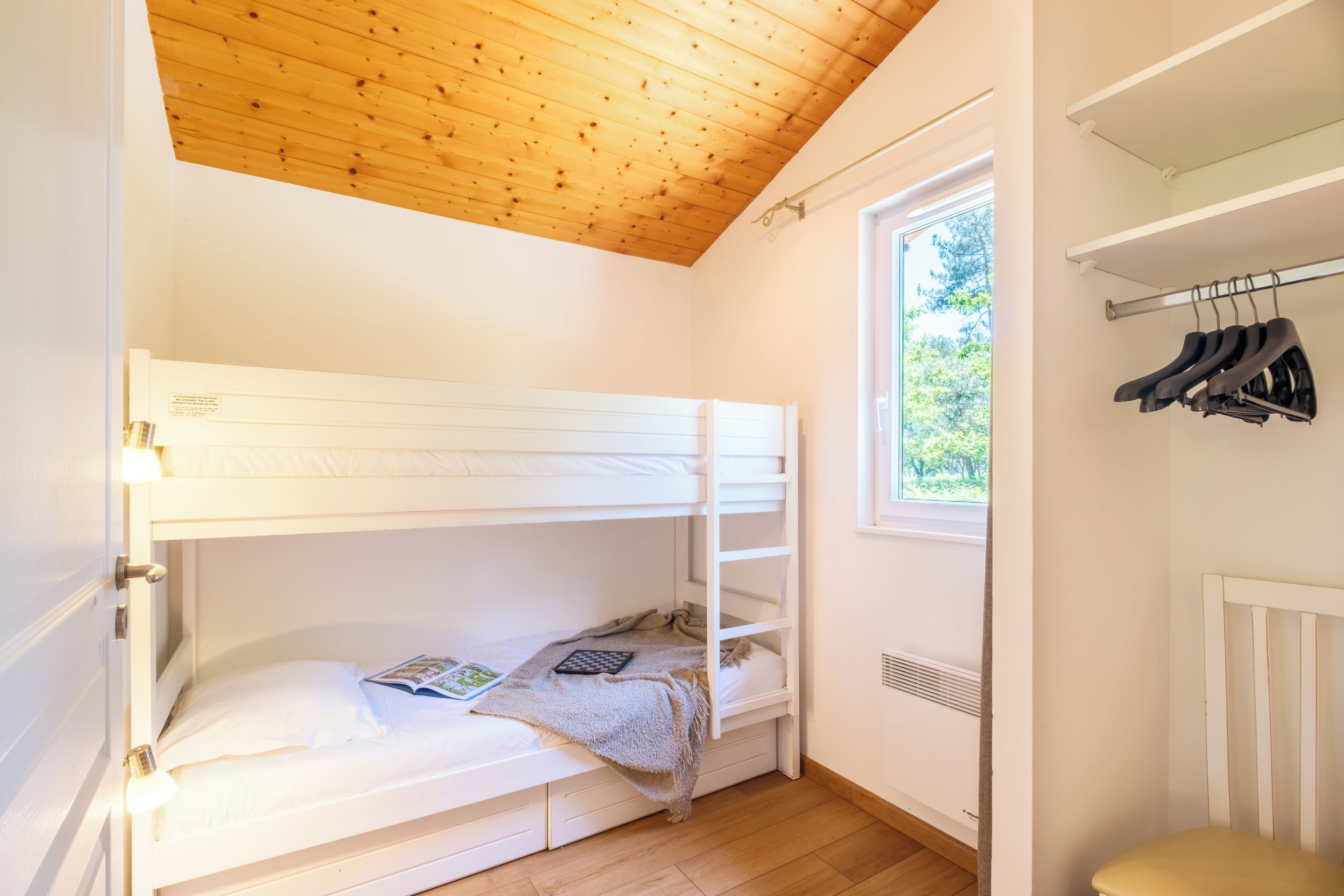 Une chambre avec un lit superposé, un plafond en bois, une fenêtre donnant sur des arbres et un radiateur.