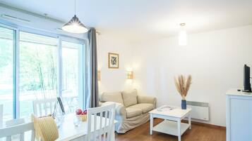 Una sala de estar moderna con un sofá blanco, una mesa de comedor con sillas, una pequeña mesa blanca y un televisor sobre un mueble.