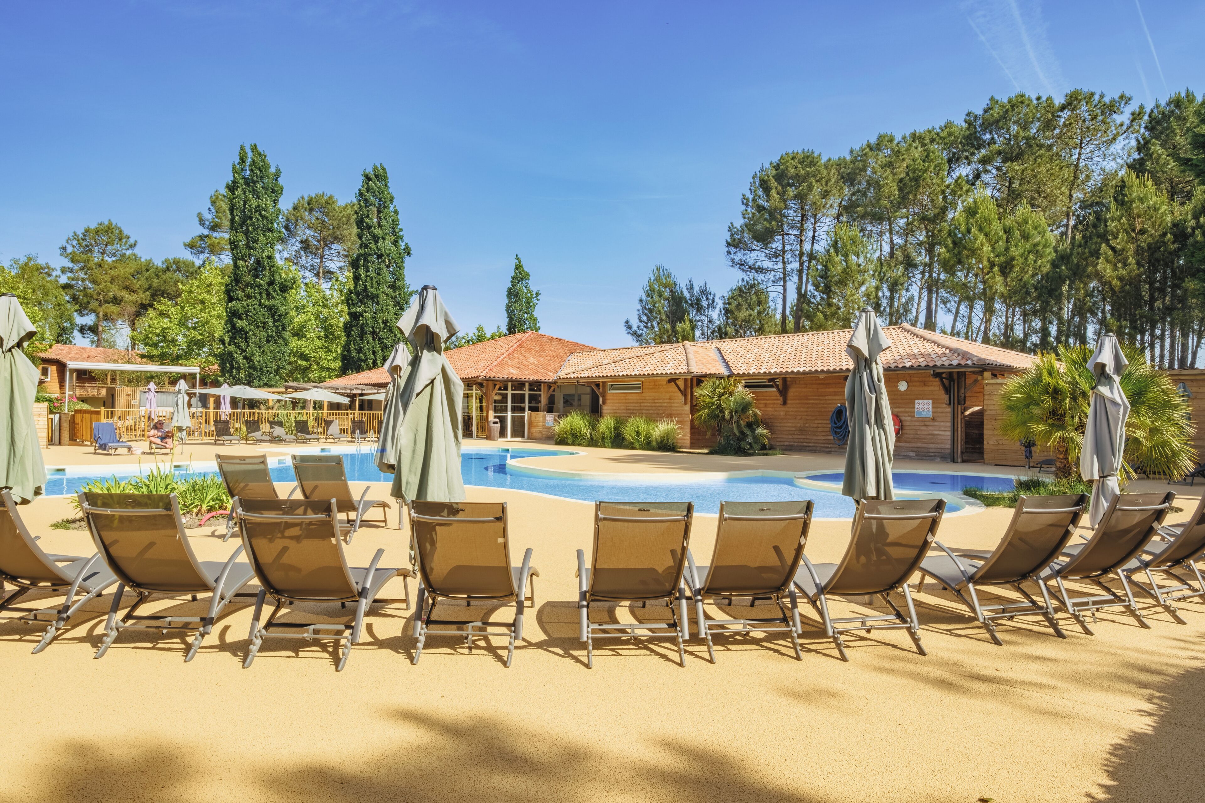 A poolside area with lounge chairs and umbrellas, surrounded by wooden buildings and tall trees.