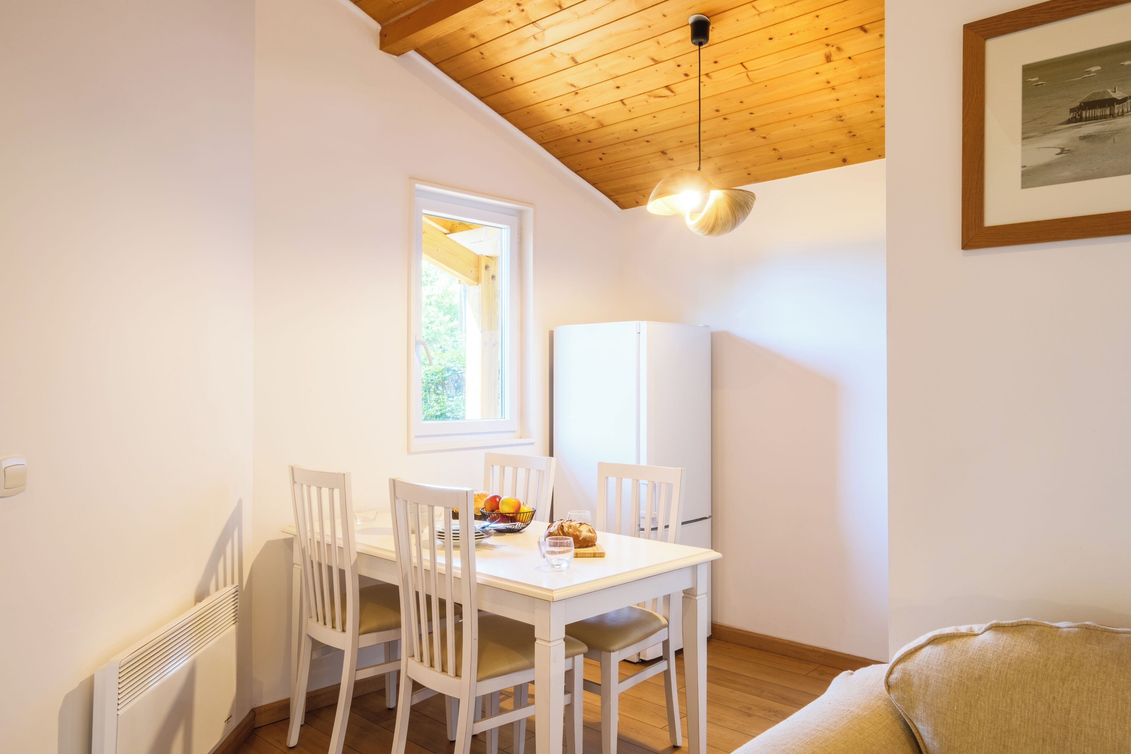 A dining area with a white table, four chairs, a window, a ceiling light fixture, and a refrigerator.