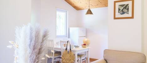 Un comedor con techo de madera, sillas blancas, una mesa con una cesta de fruta y una lámpara colgante.