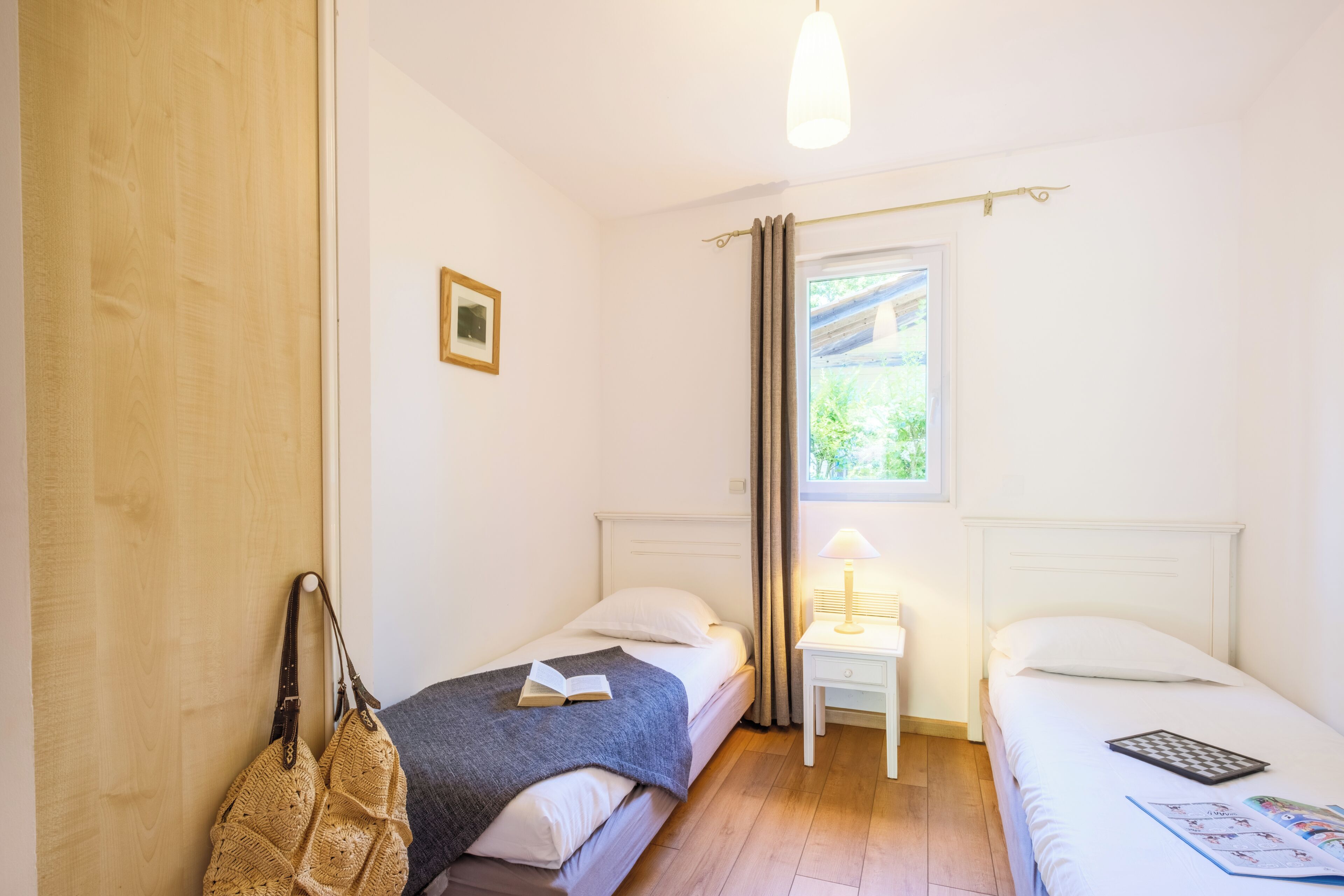 A room with two single beds, a small table with a lamp, a window with curtains, and a door.