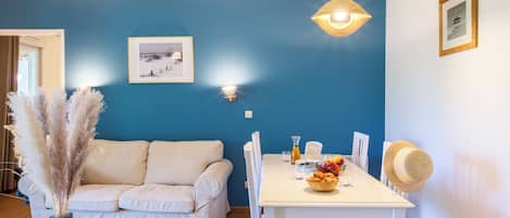 Una habitación con pared azul, sofá blanco, mesa de comedor con sillas, lámpara colgante y un cuadro enmarcado.