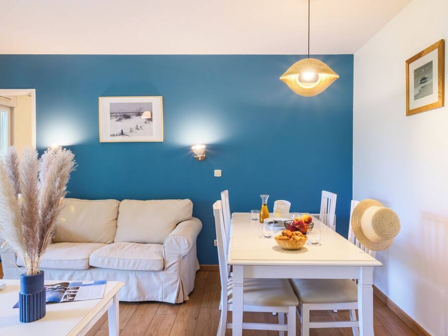 A room with a blue wall, a white sofa, a dining table with chairs, a hanging light fixture, and a framed picture.