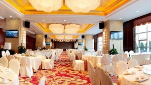 Un salón de banquetes con mesas redondas dispuestas para comer, una alfombra roja y grandes ventanales que dejan entrar luz natural.