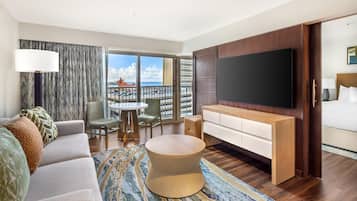 A modern hotel room with a large TV, a sofa, a round coffee table, a dining area, and a balcony with a view.
