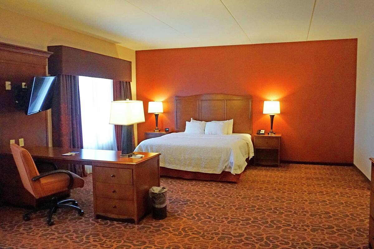 A hotel room with a bed, two bedside lamps, a desk with a chair, a TV, and a trash bin.