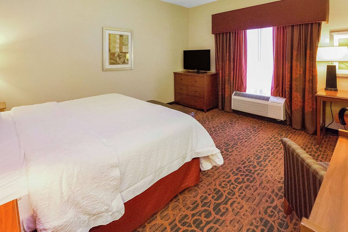 A hotel room with a bed, a television on a wooden dresser, a window with curtains, and a desk with a lamp.