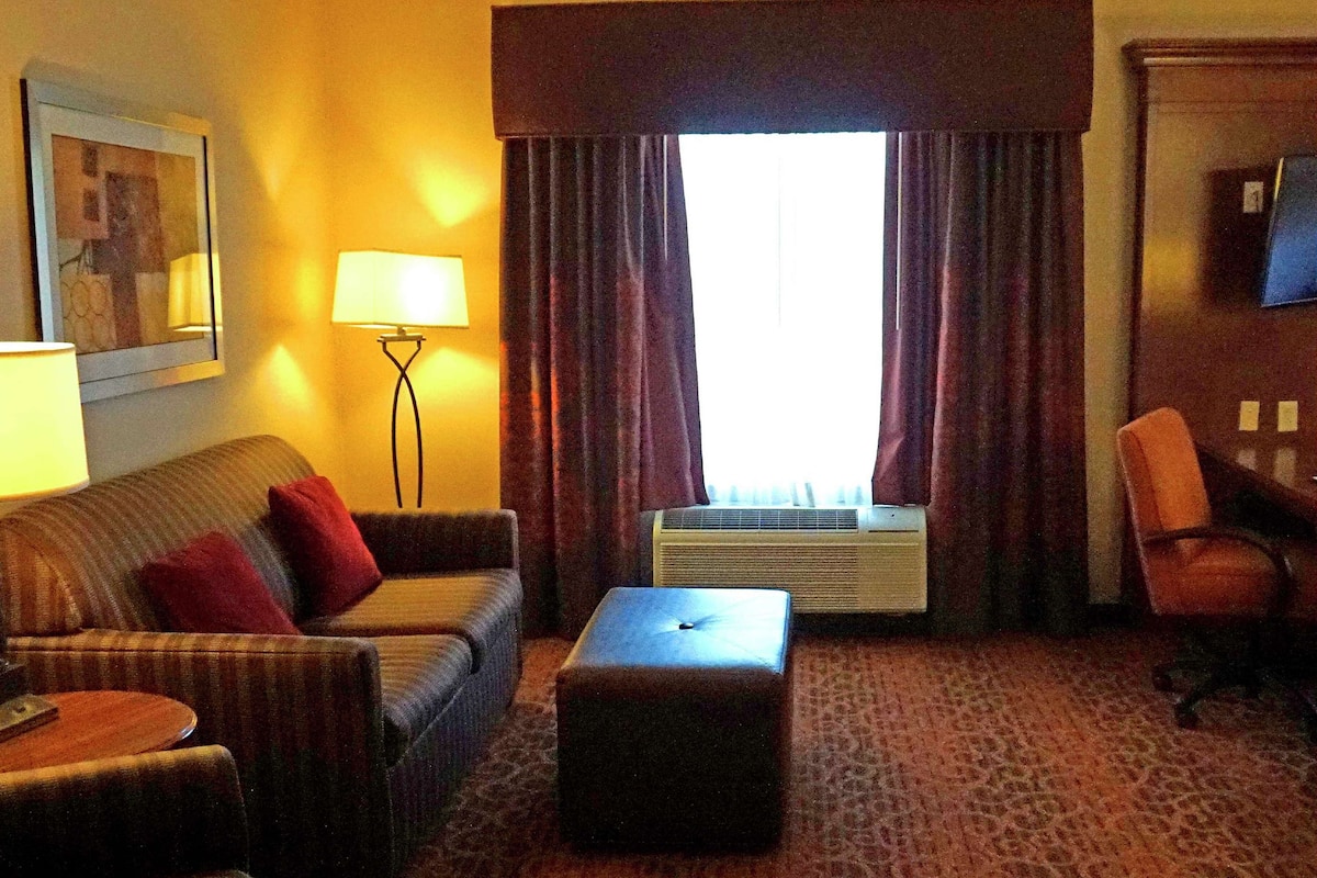 A hotel room with a sofa, ottoman, air conditioning unit, and a television on the wall.