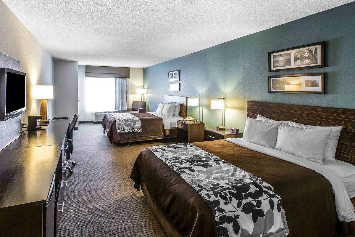 A hotel room with two beds, a desk with a flat-screen TV, and a seating area with a chair and table.