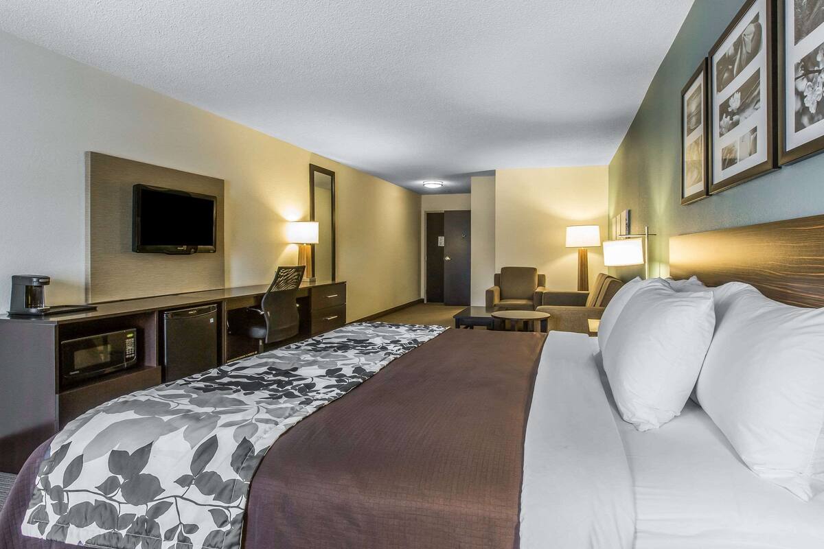 A hotel room with a large bed, a flat-screen TV mounted on the wall, a desk with a lamp, and a chair.