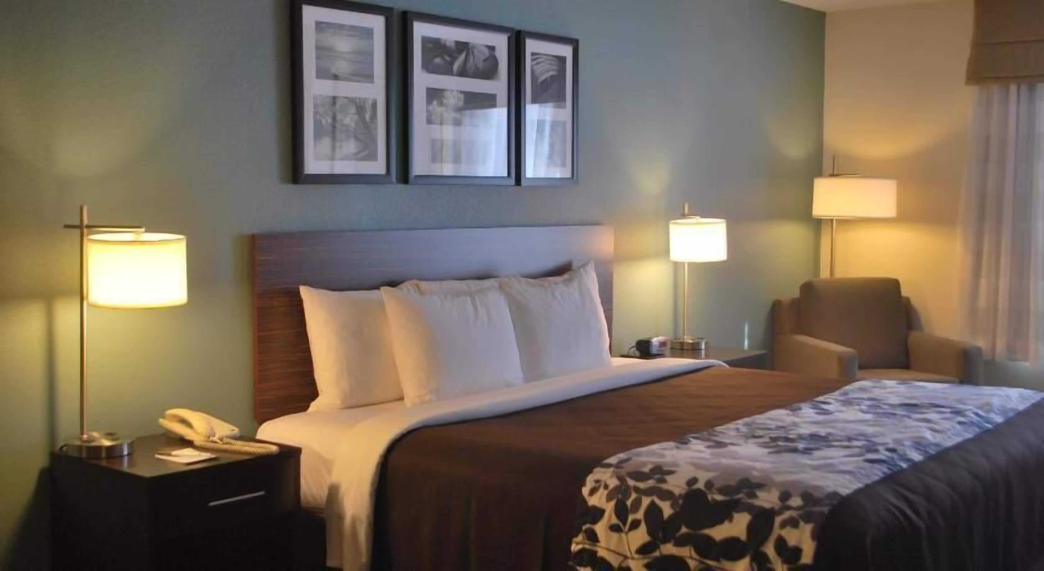A hotel room with a bed, two bedside lamps, a chair, and three framed pictures on the wall.