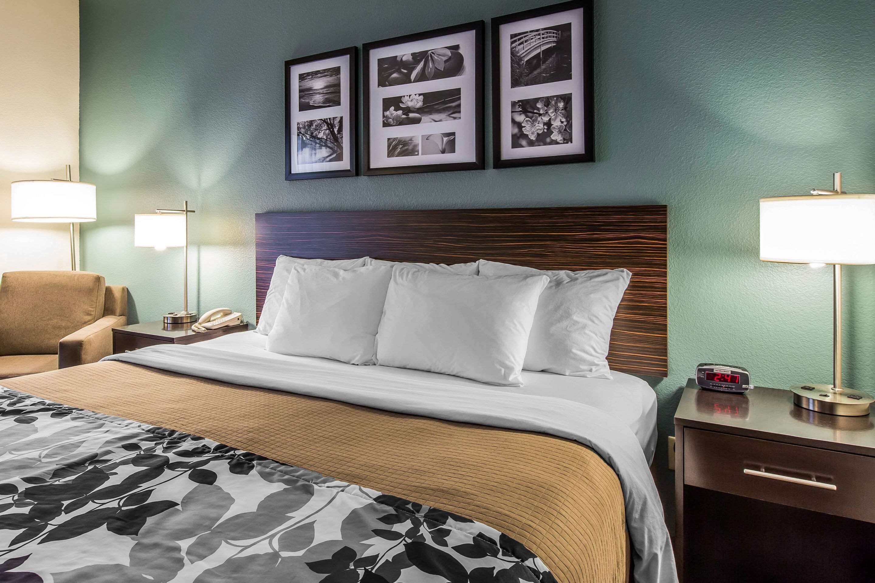 A hotel room with a large bed, a wooden headboard, a nightstand with a lamp, a chair, and framed pictures on the wall.