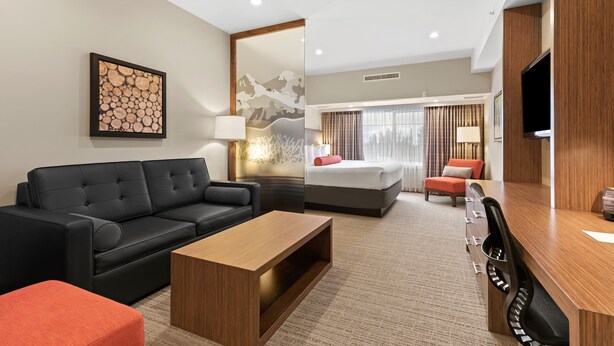 A modern hotel room with a black leather sofa, a wooden coffee table, a desk with a chair, a bed with a red pillow, and a large TV on the wall.