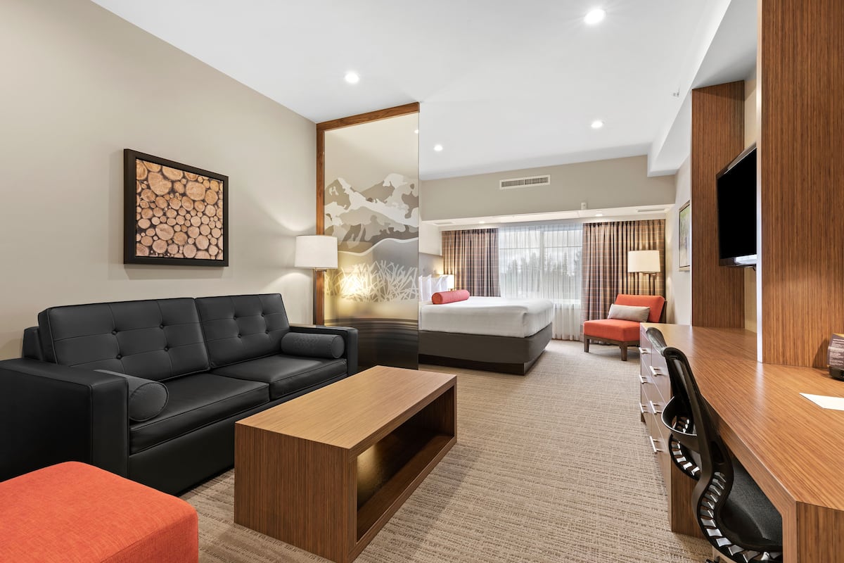 Ein modernes Hotelzimmer mit einer schwarzen Ledersofa, einem Holztisch, einem Schreibtisch mit Stuhl, einem Bett mit rotem Kopfkissen und einem großen Fernseher an der Wand.