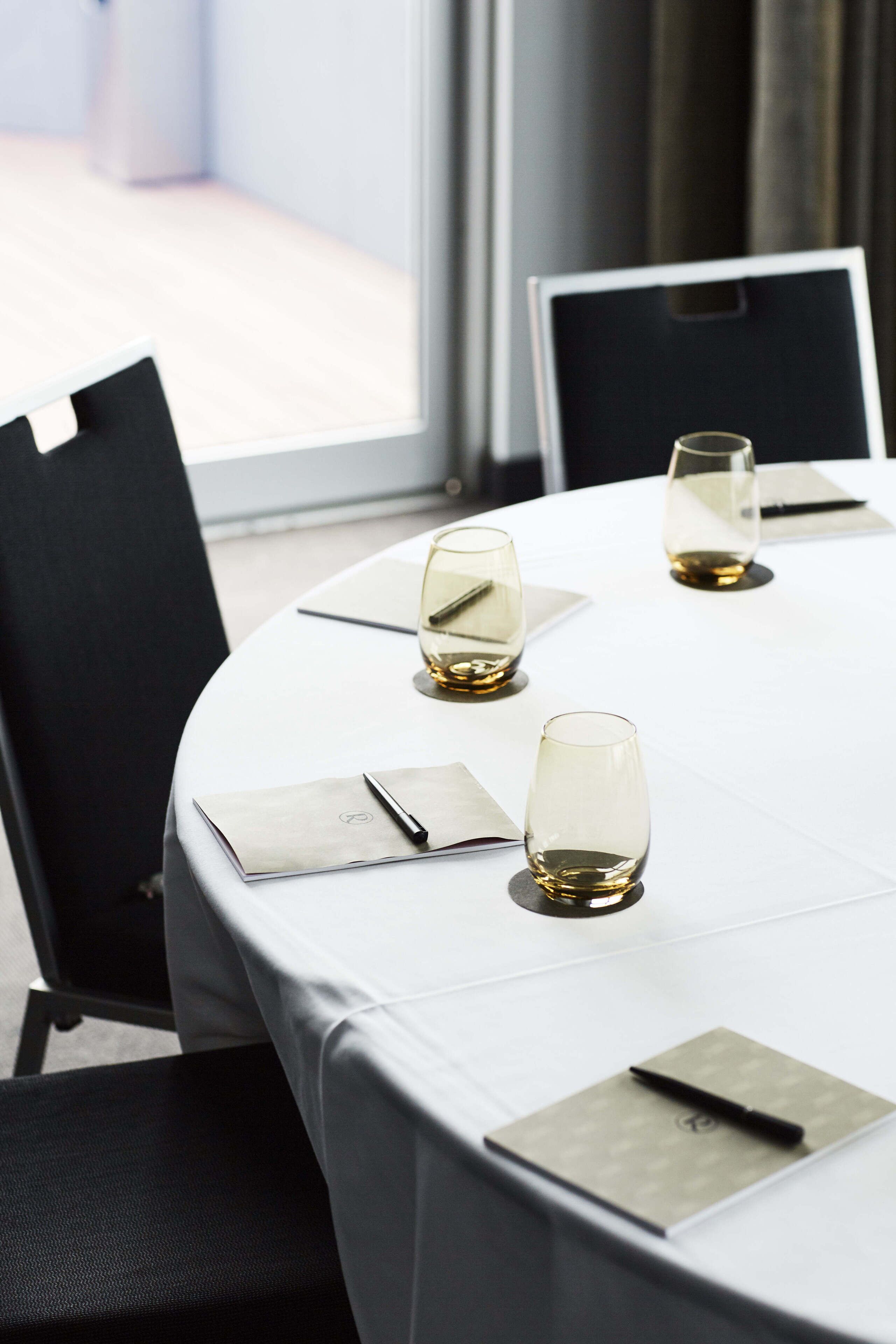 A round table with a white tablecloth, three glasses of wine, and black chairs.