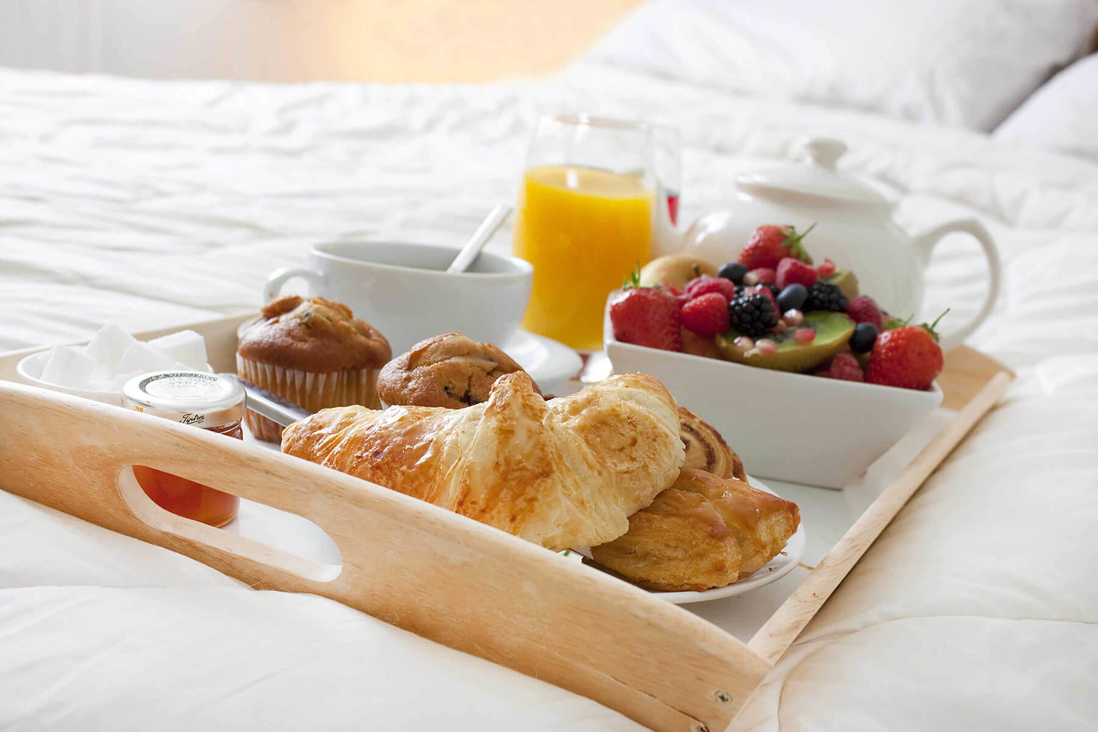 Una bandeja de desayuno con pasteles, un frutero, una jarra de jugo de naranja y una tetera sobre la cama.