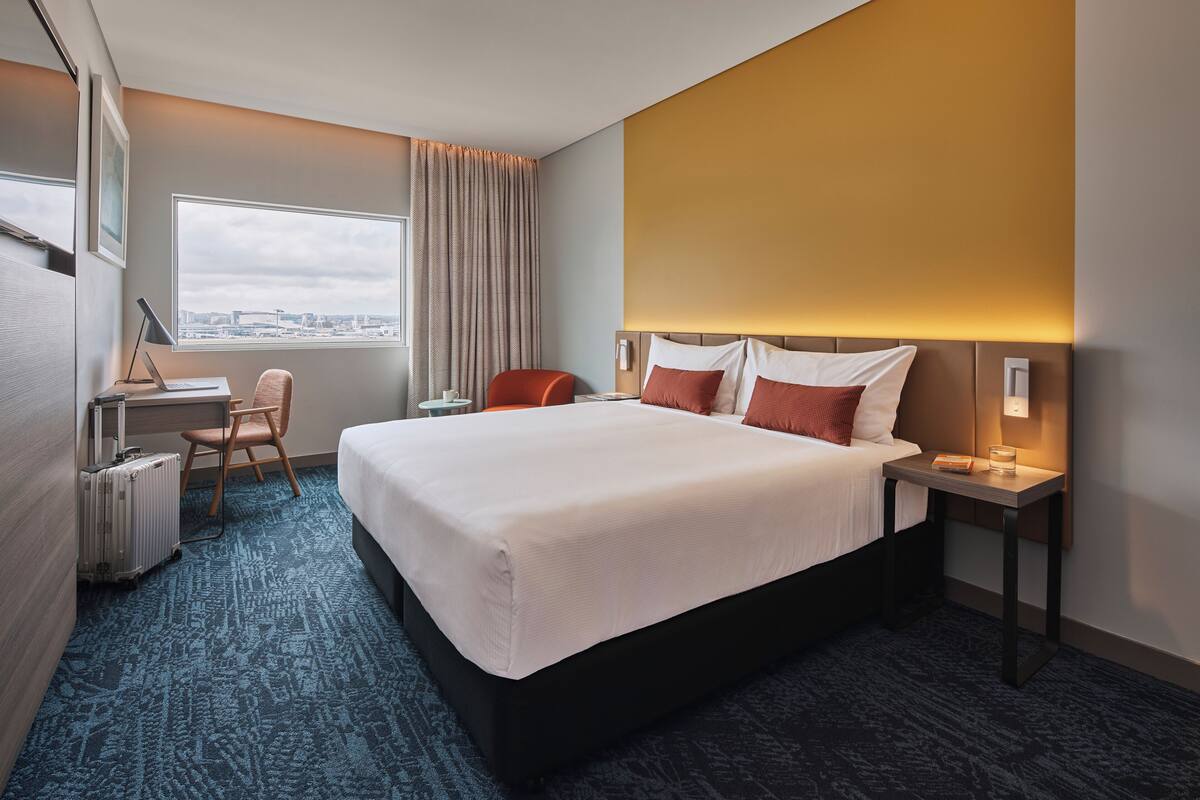 A hotel room with a large bed, a desk with a laptop, a chair, a suitcase, and a view of the city through a large window.