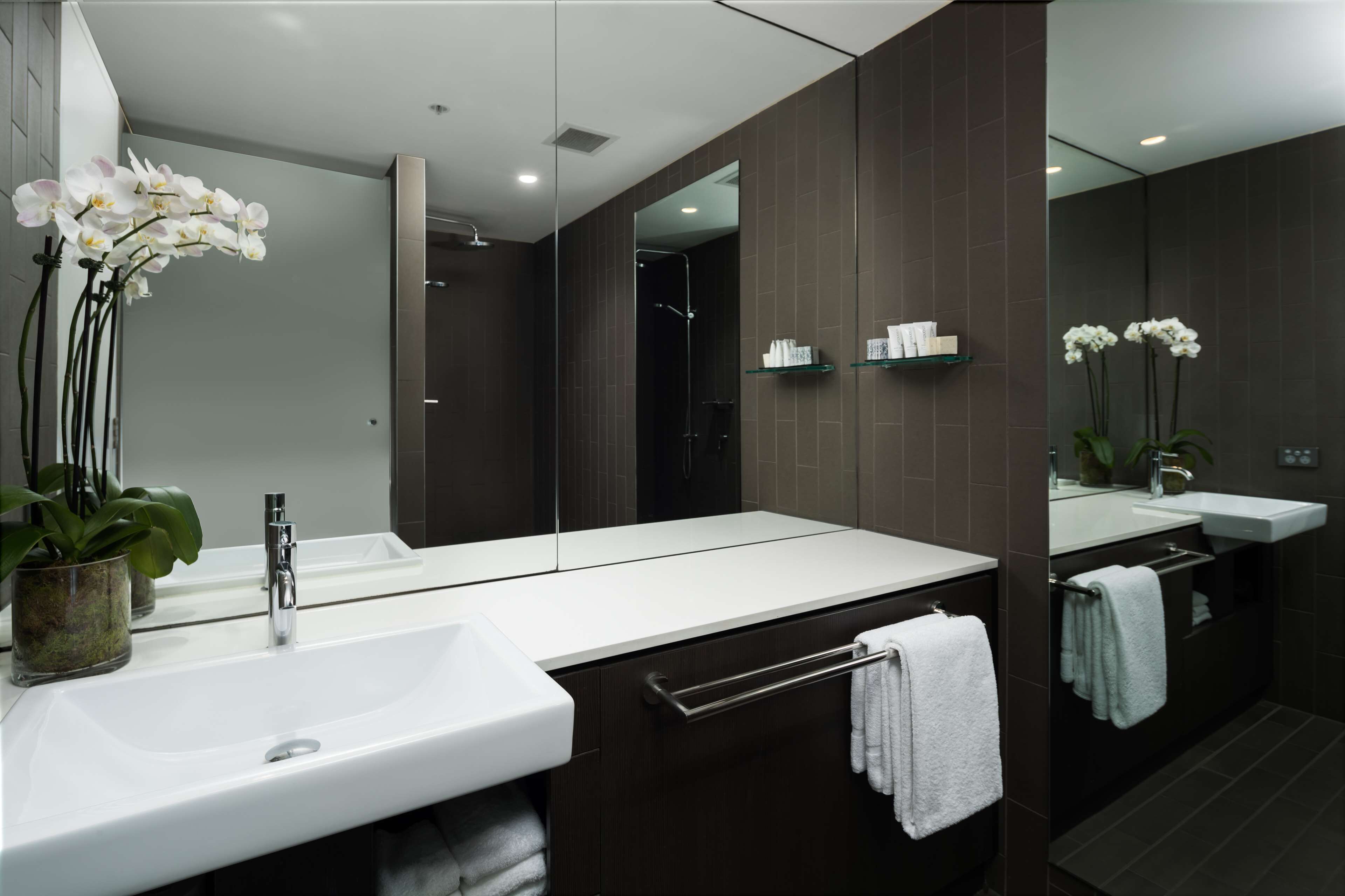 A modern bathroom with a large mirror, white sink, and dark brown tiles. There are white orchids on the left and a towel rack with white towels.