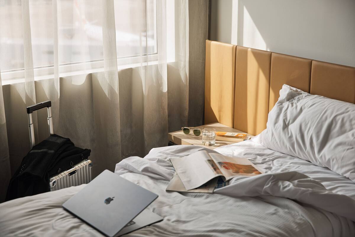 A hotel room with a bed, a suitcase, a laptop, a magazine, and a glass of water on a nightstand.