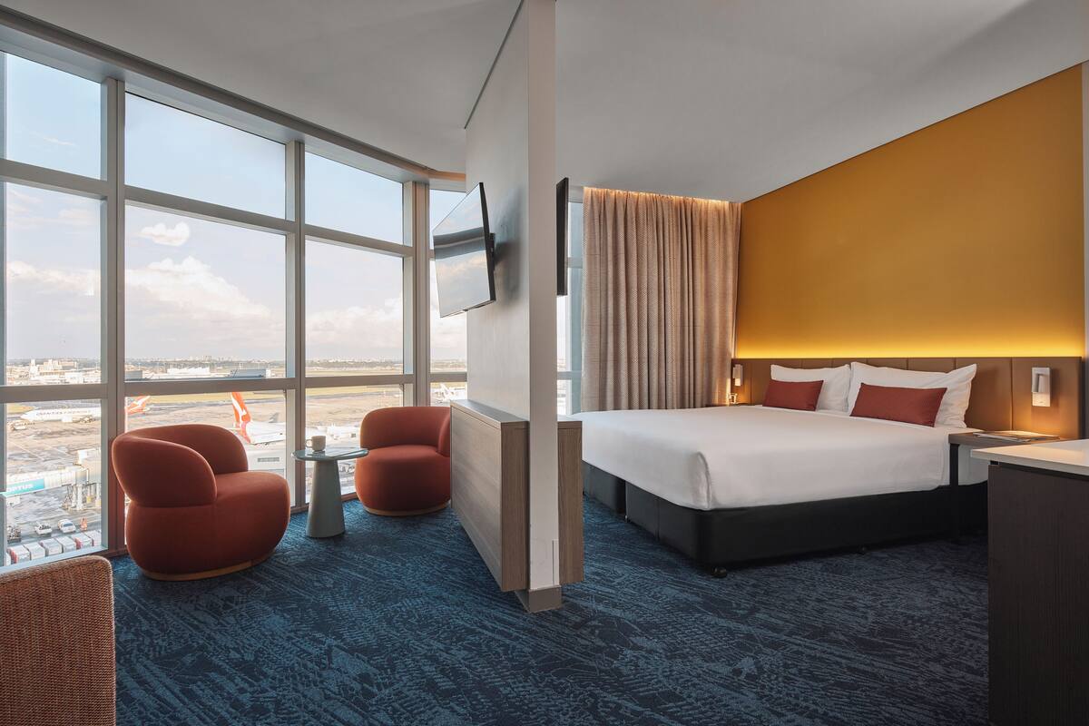 A hotel room with a large window, a bed, a TV, and a seating area with two chairs.