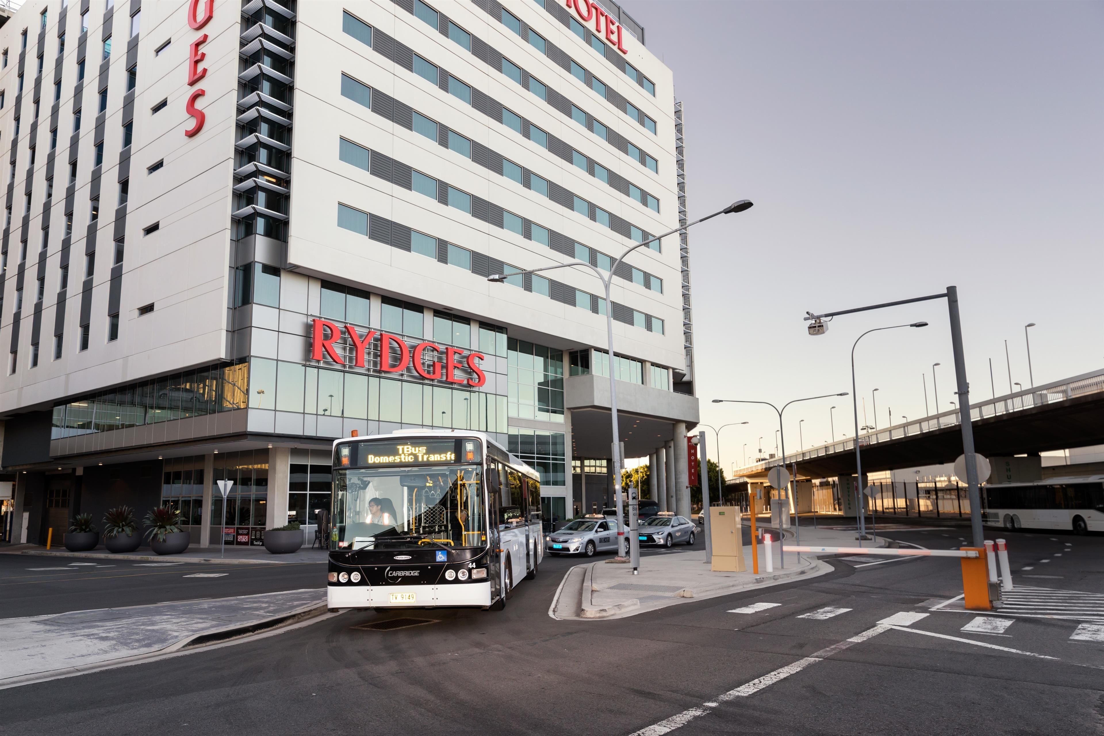 A modern hotel with a prominent 'RYDGES' sign, a bus in front, and a pedestrian crossing.