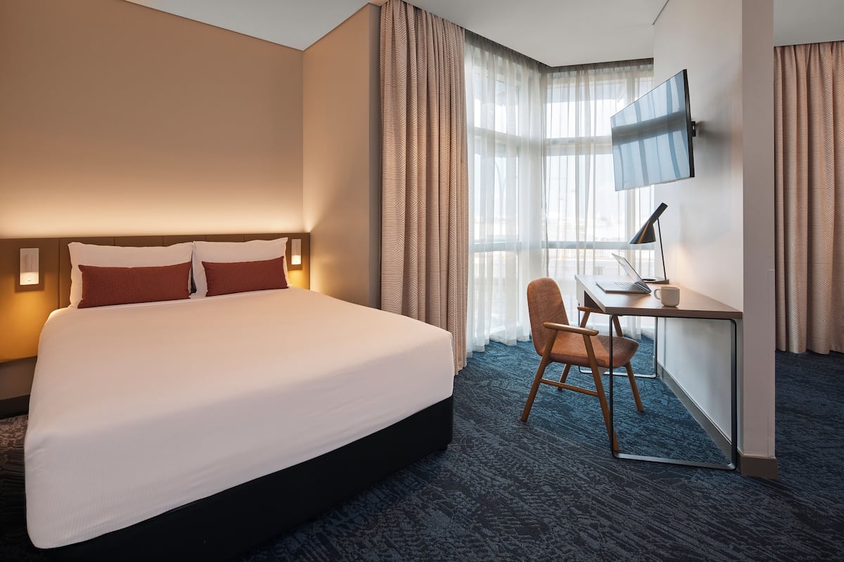 A hotel room with a large bed, a desk with a chair, a flat-screen TV, and a window with curtains.