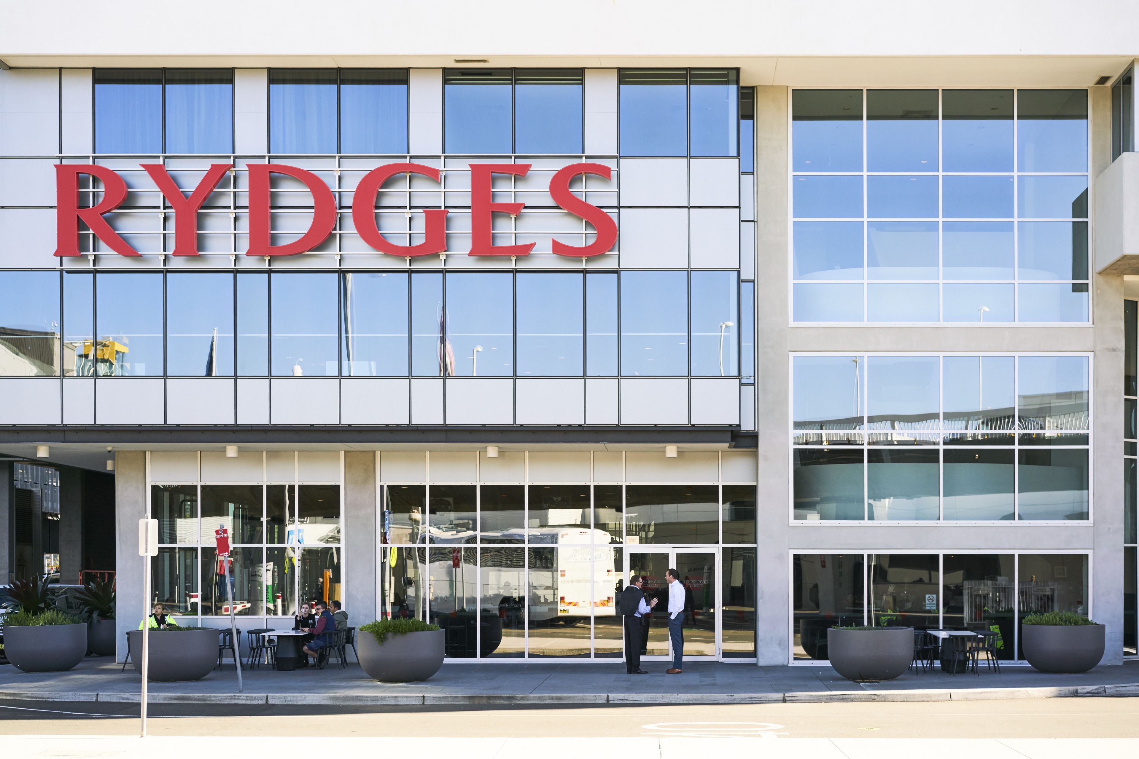 A Rydges hotel with large windows, outdoor seating, and potted plants.
