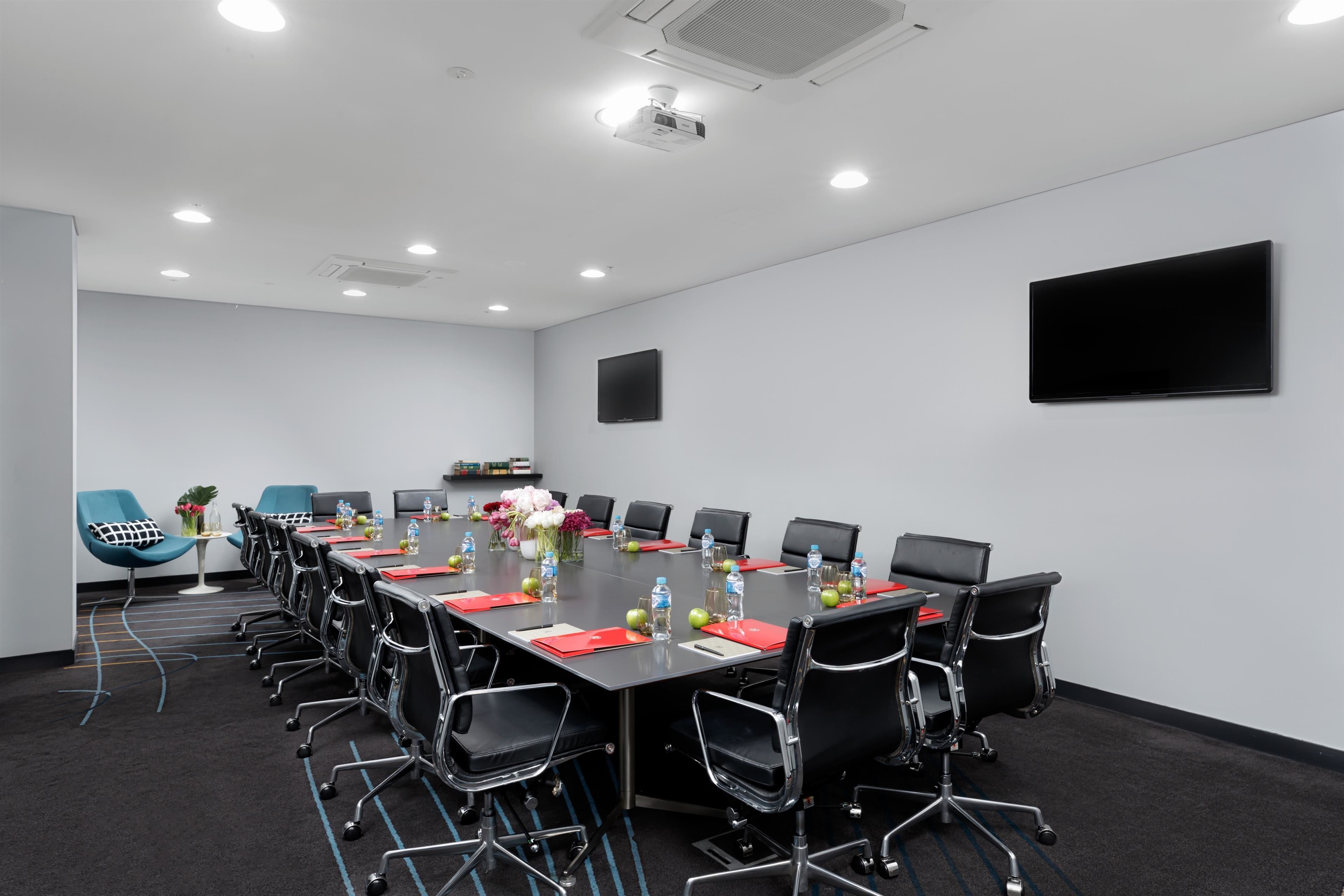 Uma sala de conferências com uma mesa comprida, cadeiras pretas e uma televisão de tela plana na parede.