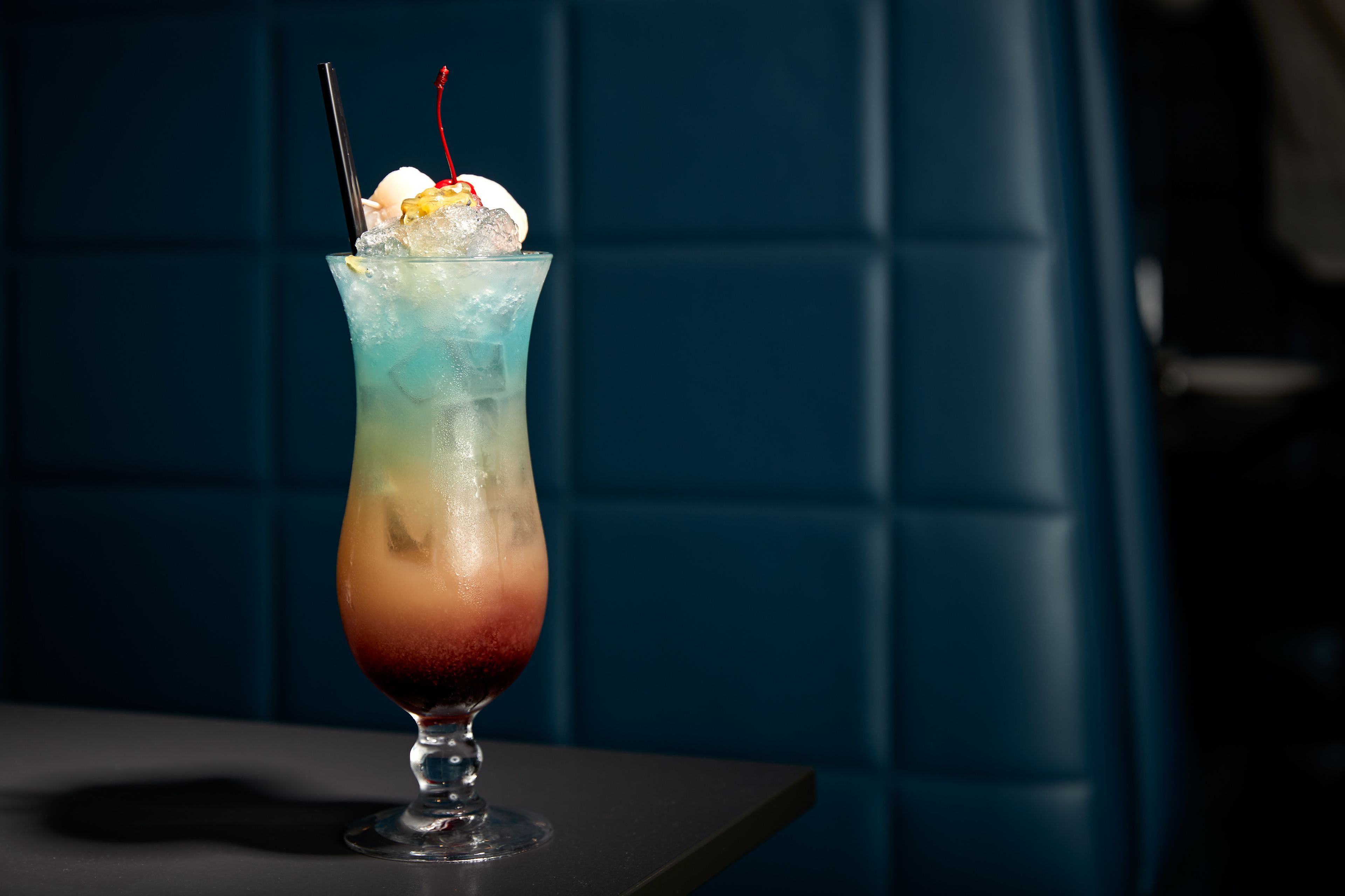 Um cocktail num copo alto com efeito degradê de cores, cubos de gelo e uma cereja por cima.