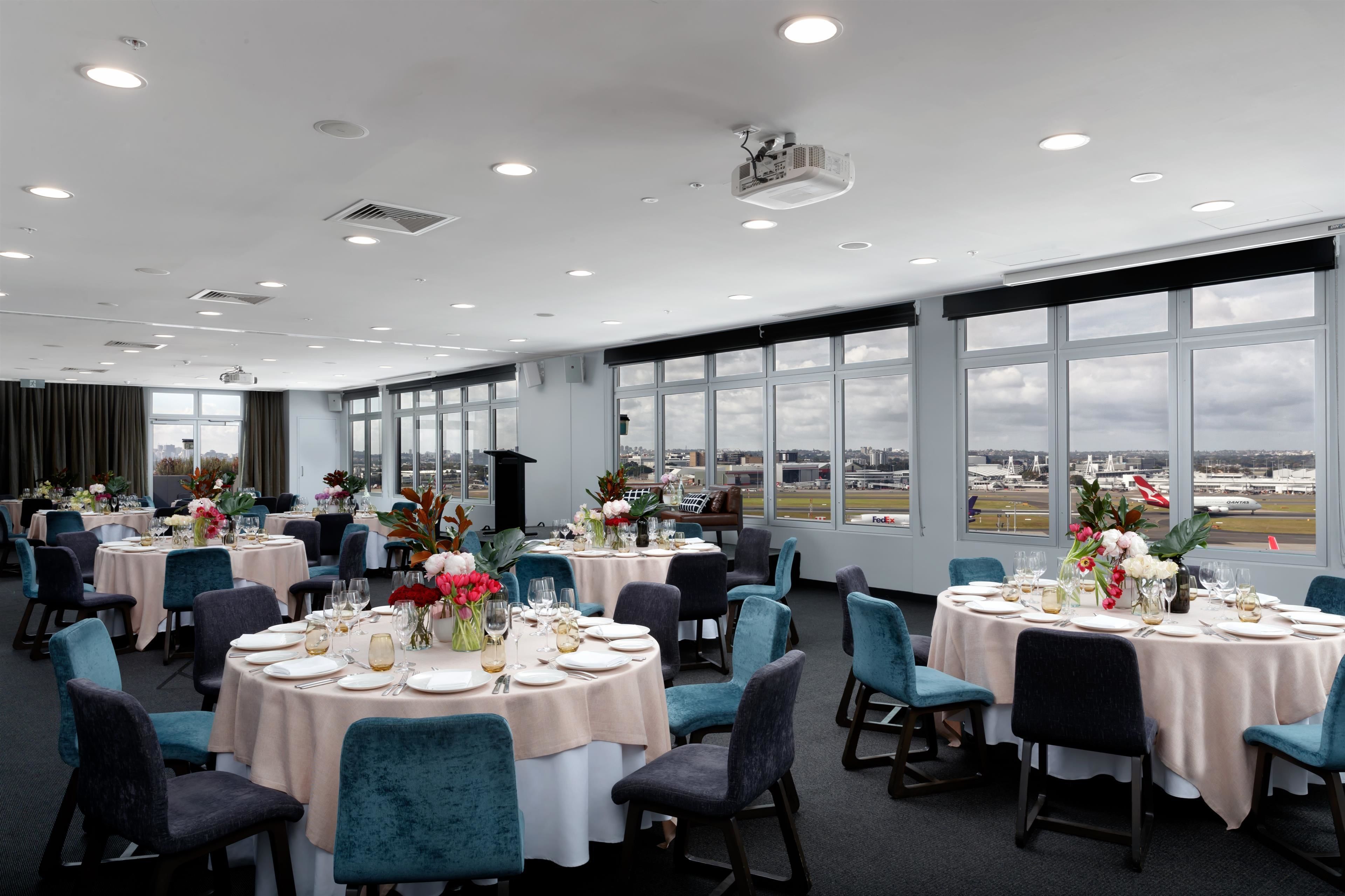 A spacious dining area with round tables, teal chairs, and large windows offering a view of the airport.