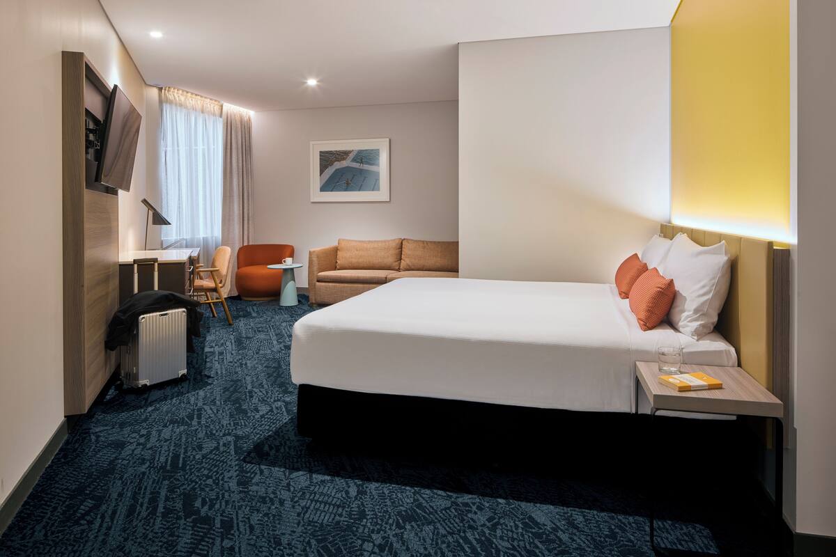 A hotel room with a bed, a desk, a chair, a radiator, and a window with curtains.