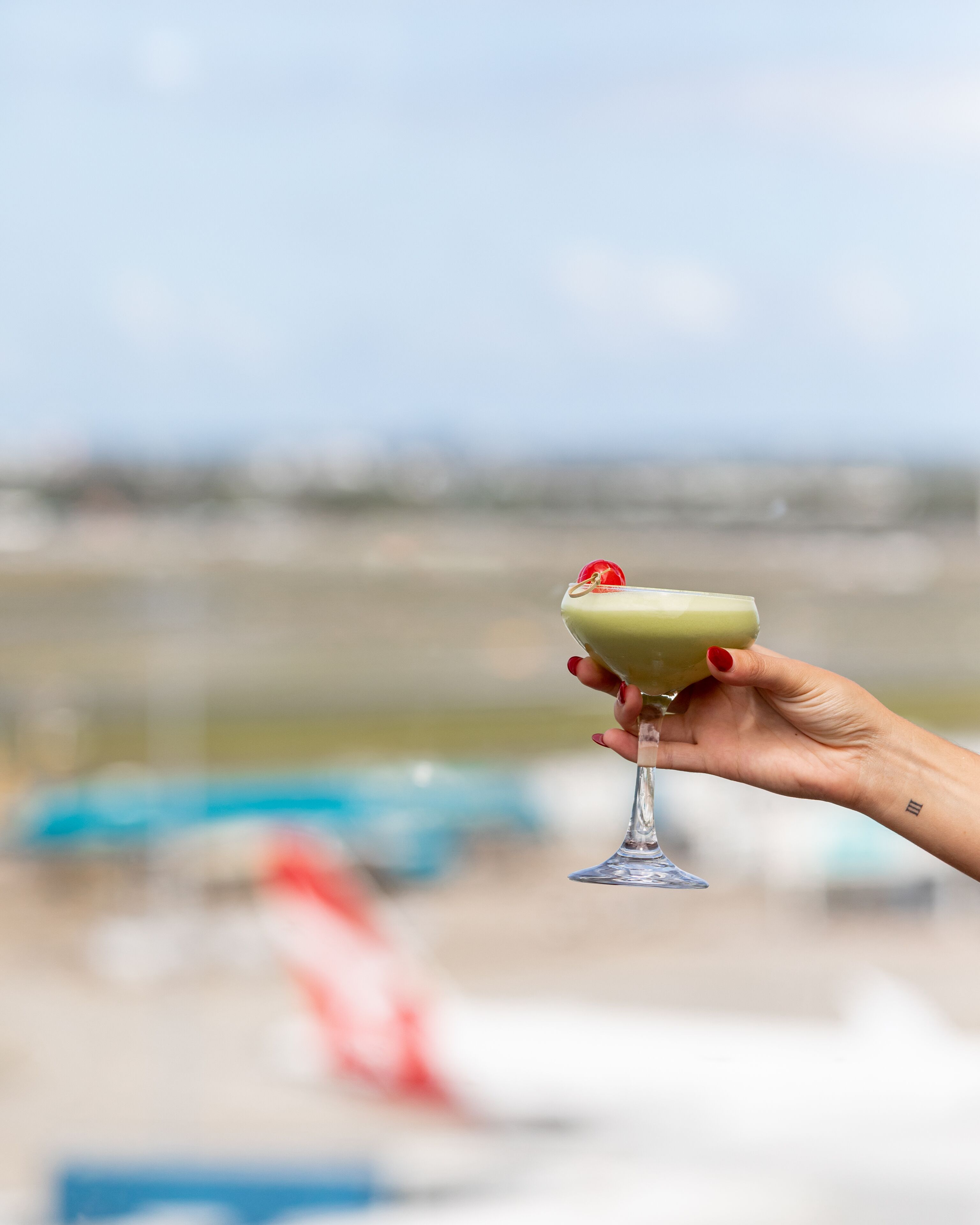 A hand holding a cocktail glass with a green drink and a cherry garnish, against a blurred background with a lifeguard tower.