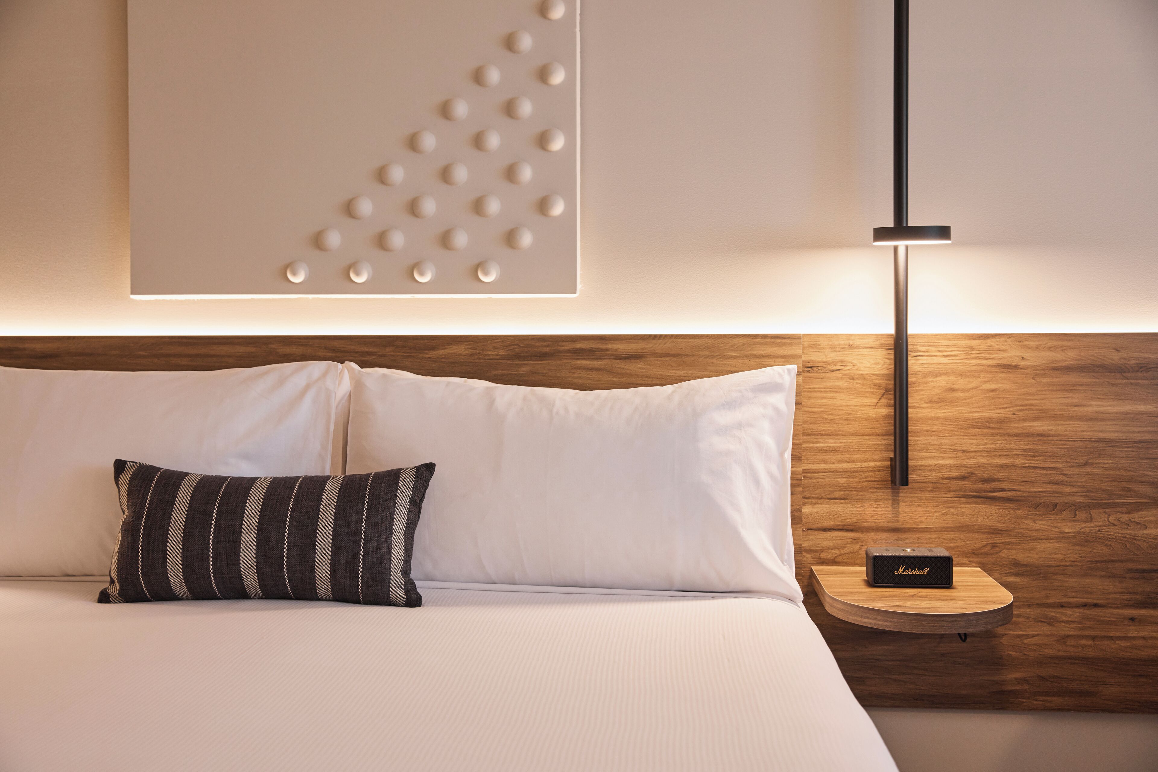 Een net opgemaakt bed met witte lakens en een gestreept kussen, een wandlamp en een klein houten nachtkastje met een speaker.