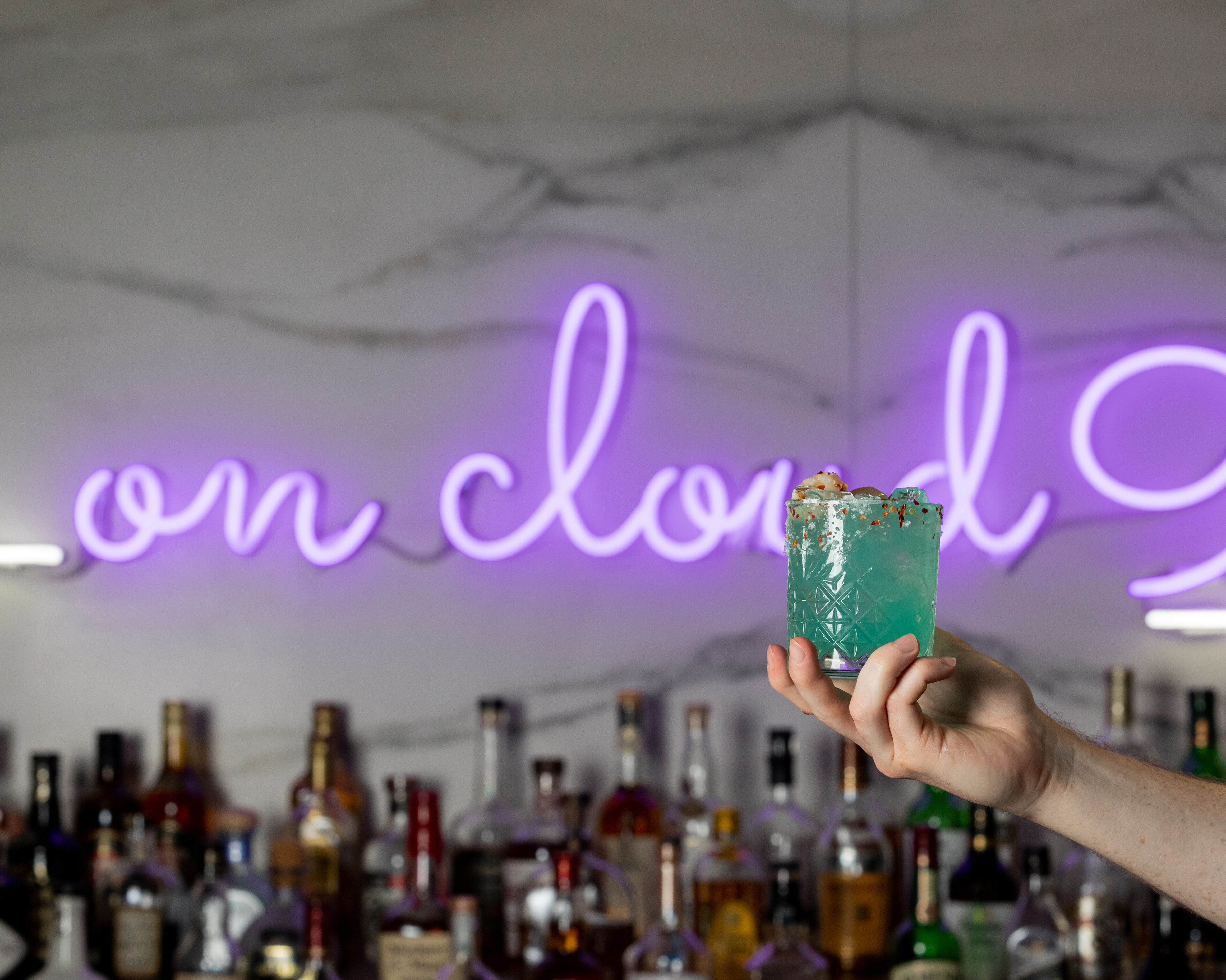 A hand holding a green cocktail with ice cubes and garnishes, in front of a neon sign that reads "on cloud nine."