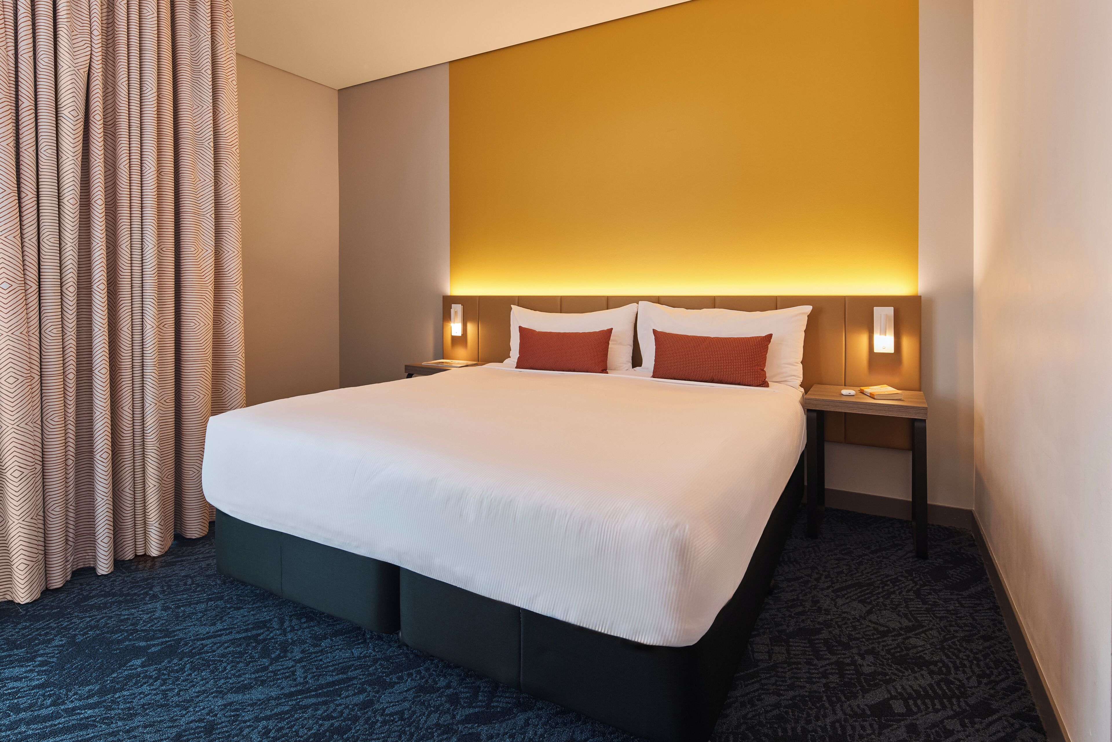 Ein Hotelzimmer mit einem großen Bett, zwei Nachttischen, einer Wandlampe und einem gemusterten Vorhang.