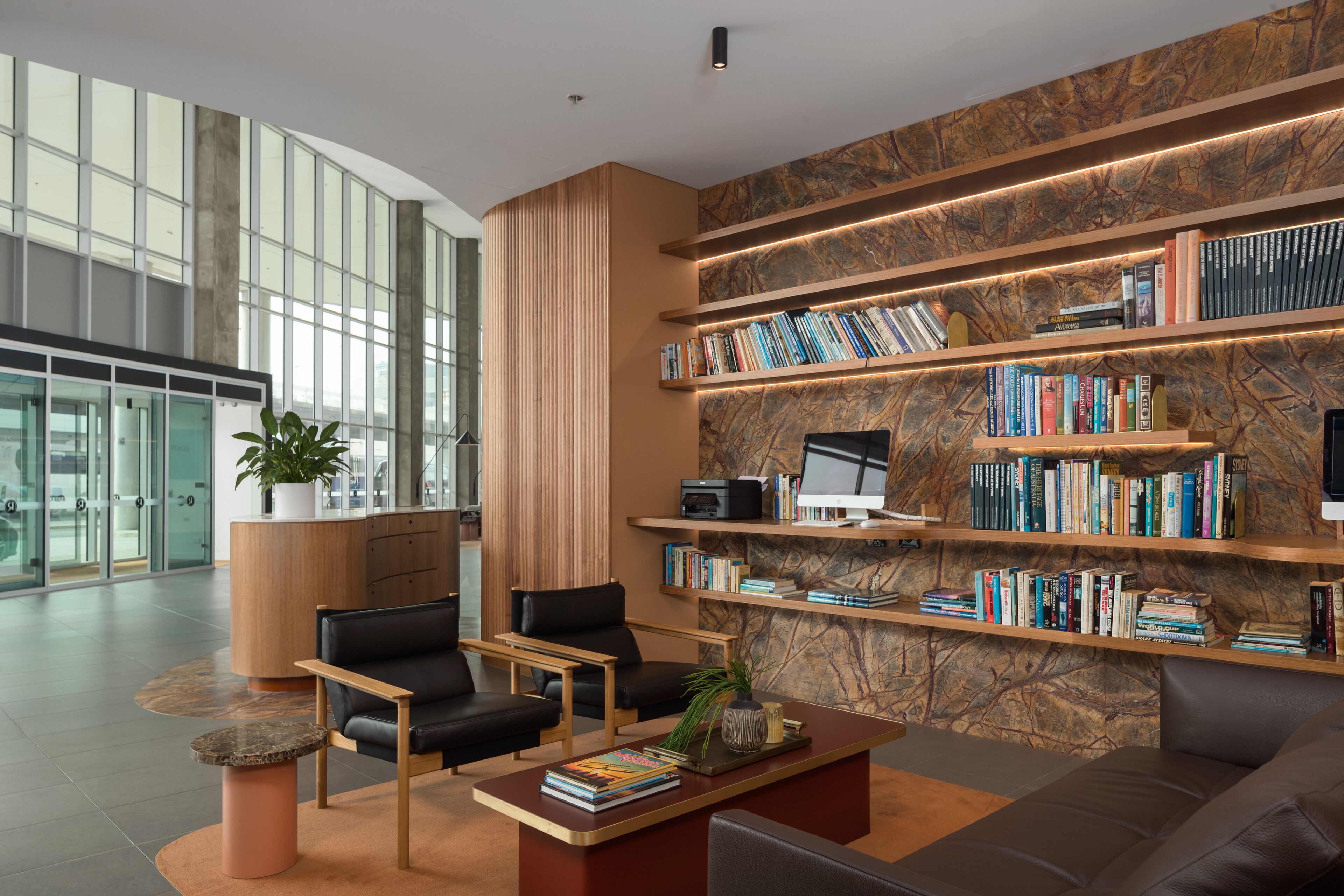 A modern lobby with a curved glass wall, a central seating area with a coffee table, and a marble wall with bookshelves.