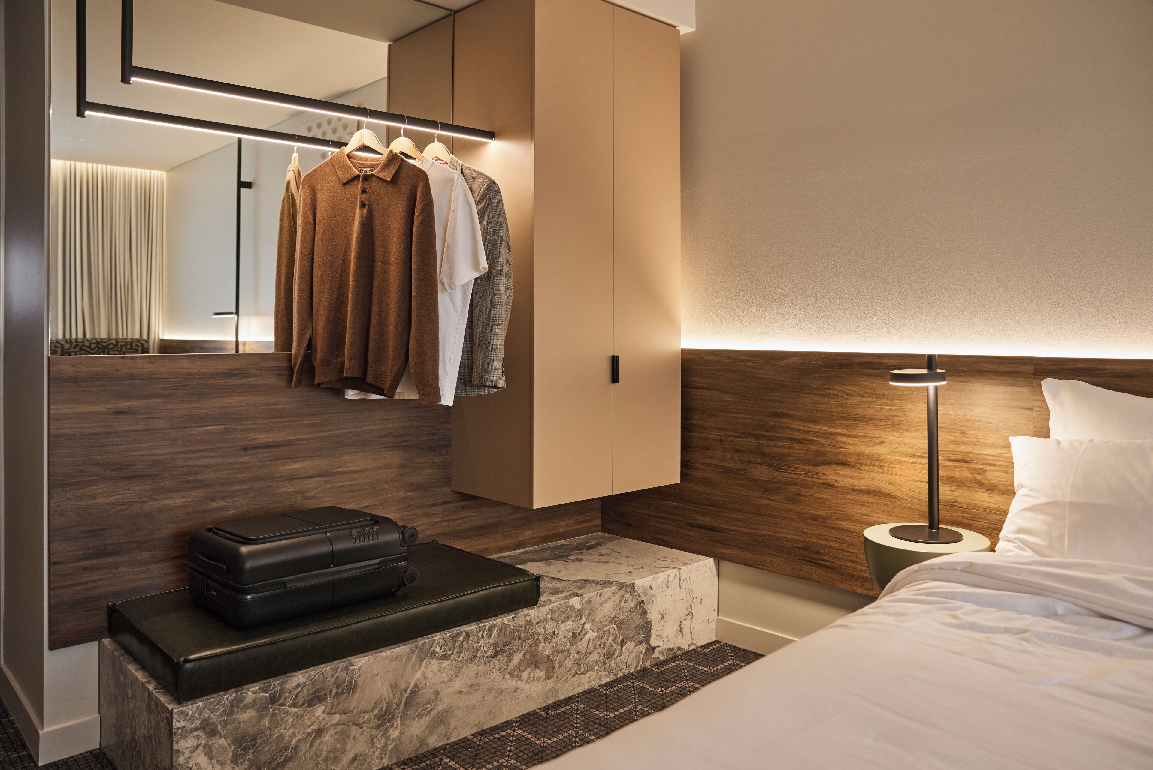 Una habitación de hotel moderna con cabecera de madera, una mesita de noche con una maleta, armario ropero y un espejo grande.
