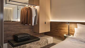 Una habitación de hotel moderna con cabecera de madera, una mesita de noche con una maleta, armario ropero y un espejo grande.