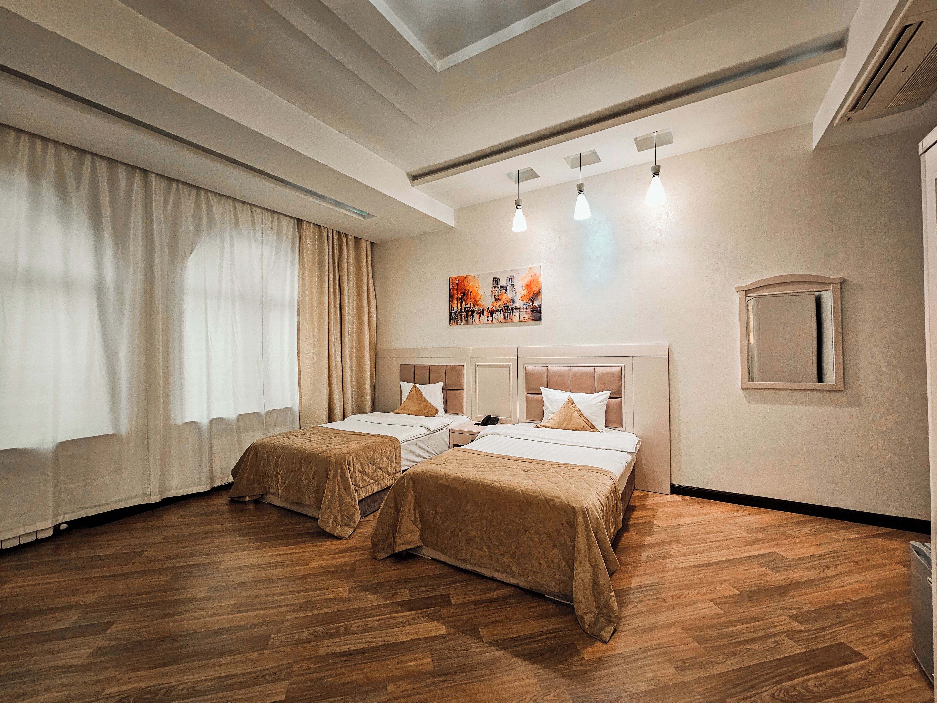 Une chambre d’hôtel avec deux lits, un plancher en bois, une grande fenêtre avec des rideaux, un tableau au mur et un luminaire au plafond.