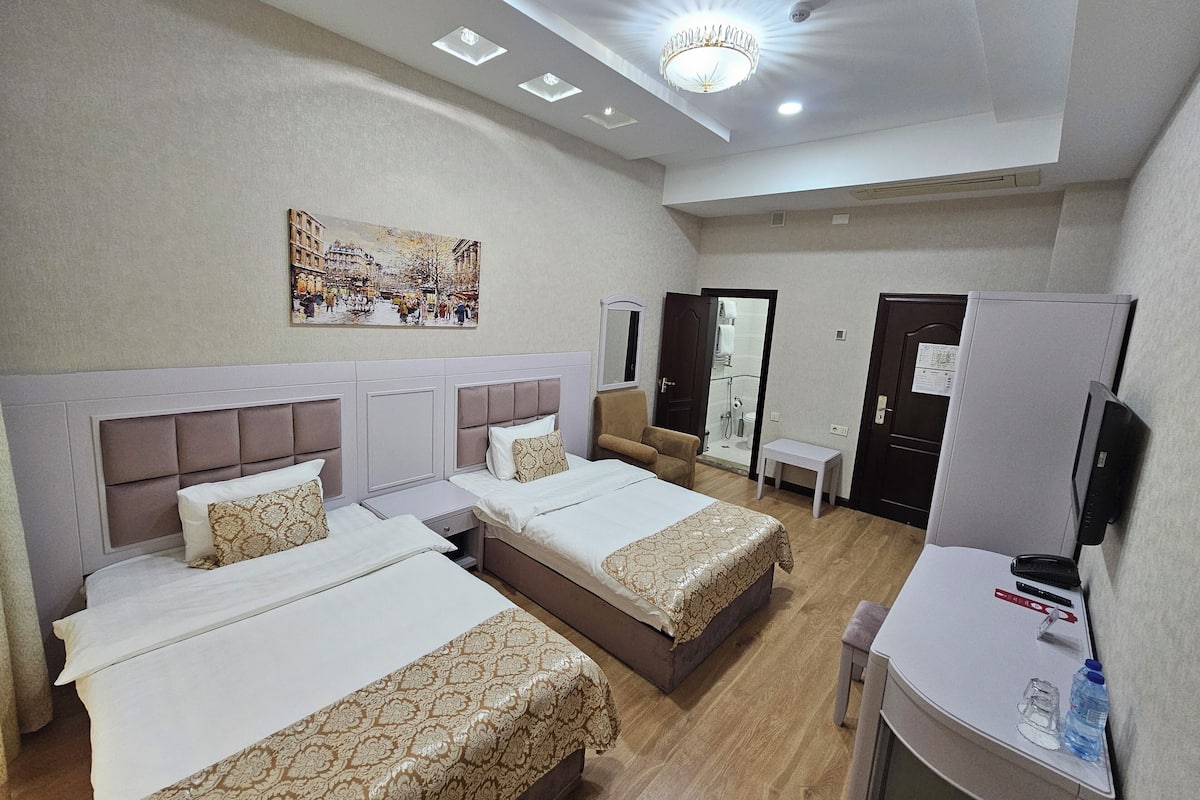 A hotel room with two beds, a small table, a mirror, a wardrobe, and a shower area.