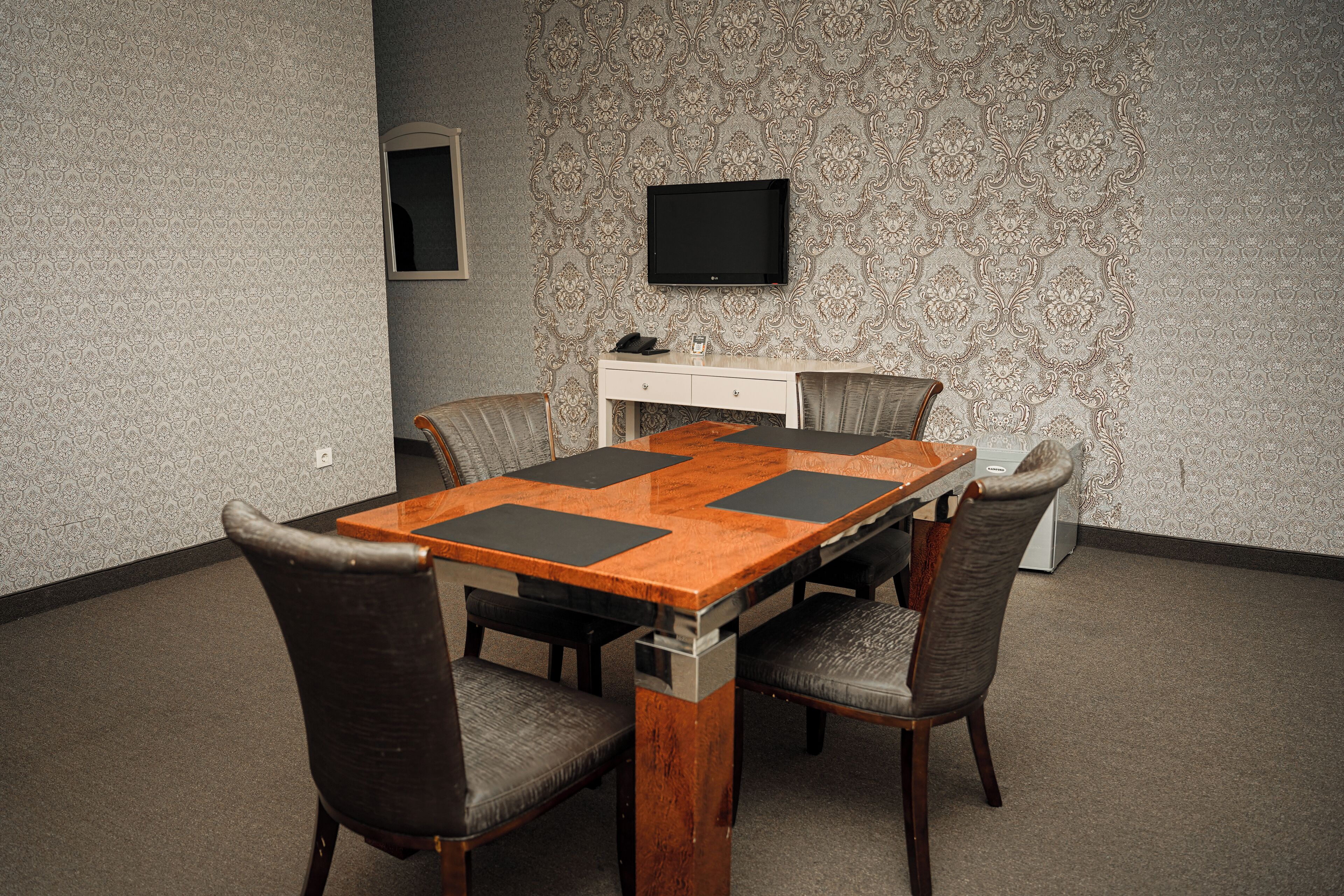 Un salón de conferencias con una mesa de madera, cuatro sillas, un escritorio blanco, un televisor de pantalla plana y papel tapiz estampado.