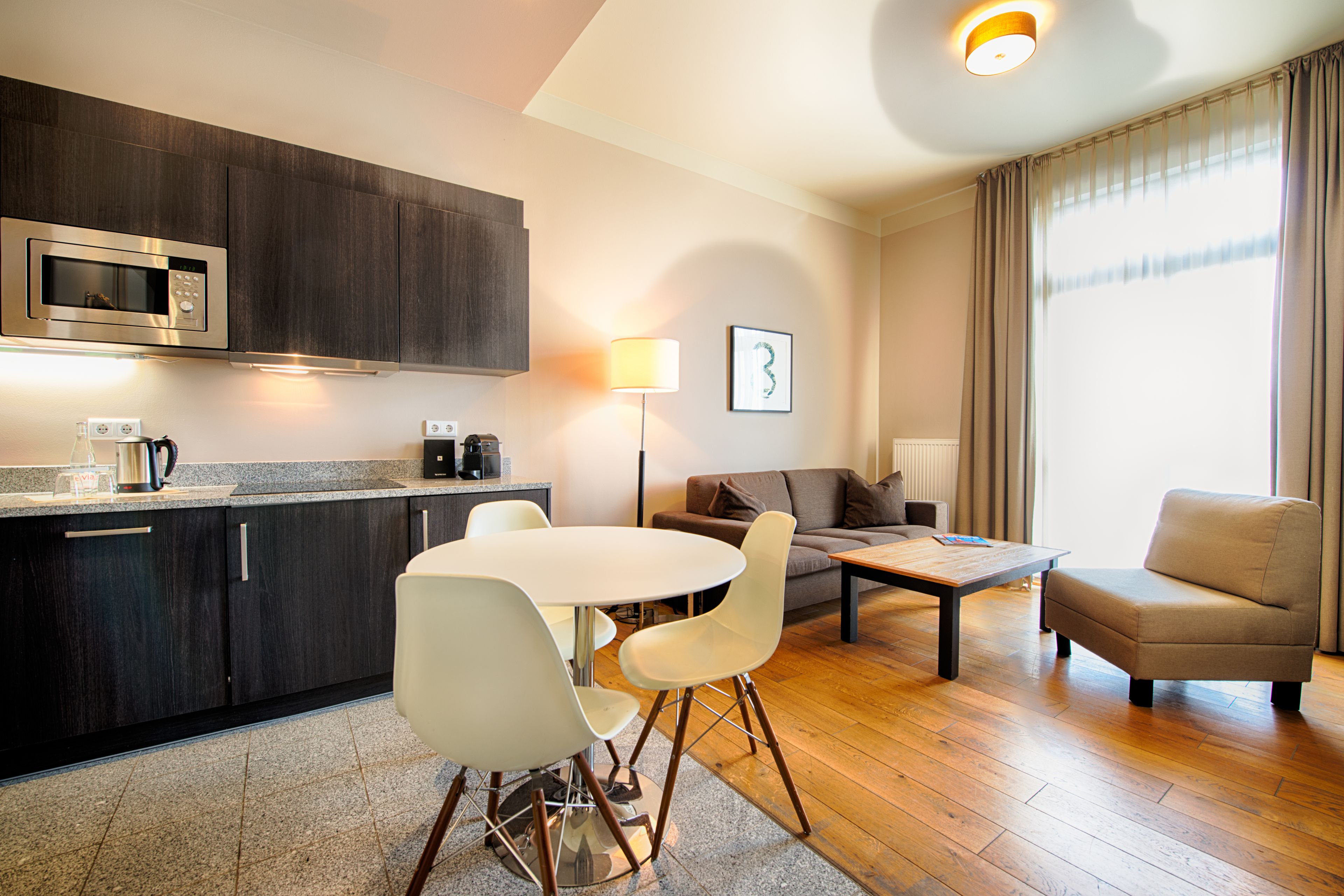 A modern kitchen with dark cabinets, a microwave, and a coffee maker. There is a dining area with a white table and chairs, a sofa, and a lamp. The room has wooden flooring and a large window with curtains.