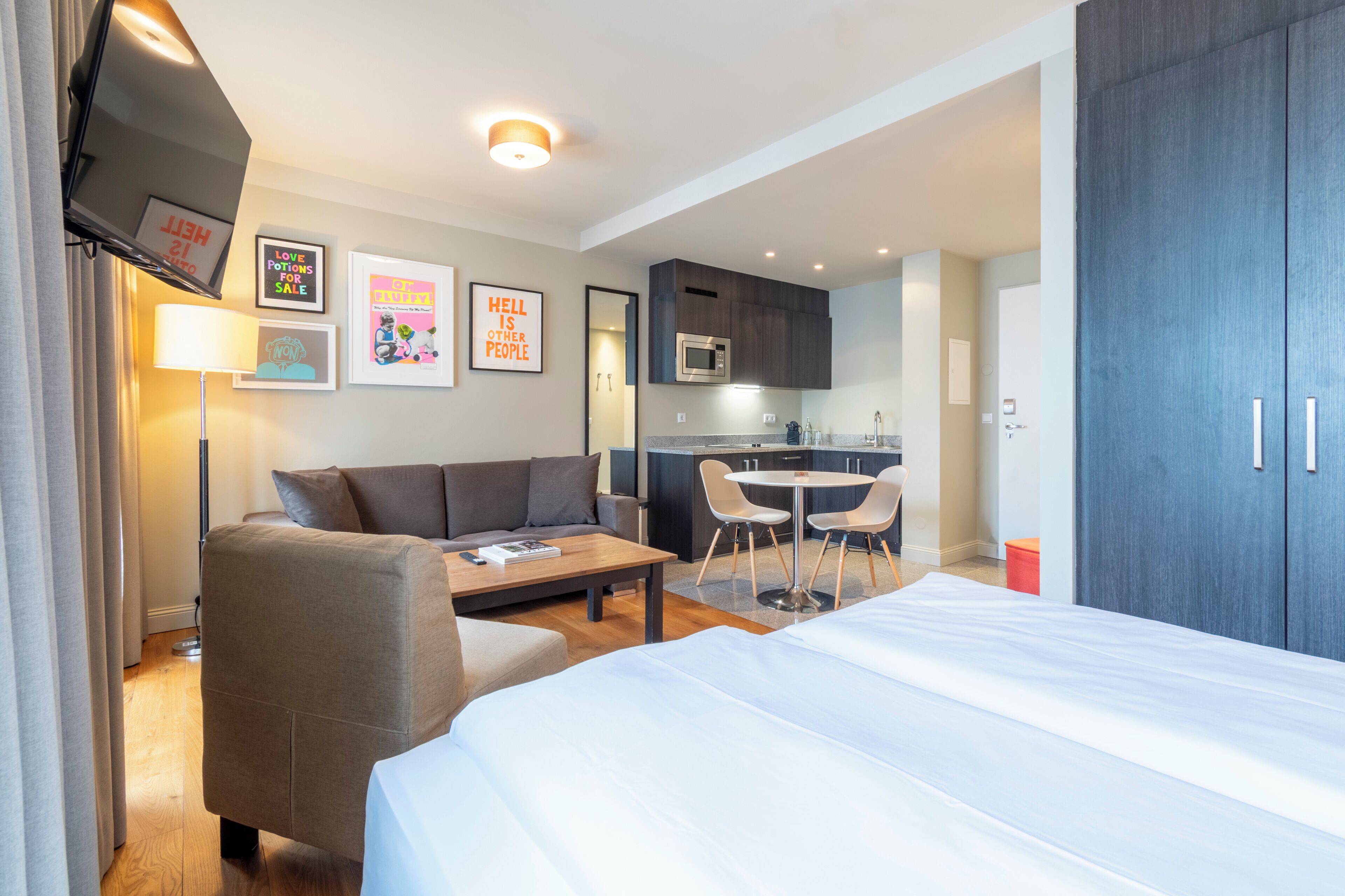 A modern hotel room with a bed, a sofa, a dining table, and a kitchenette.