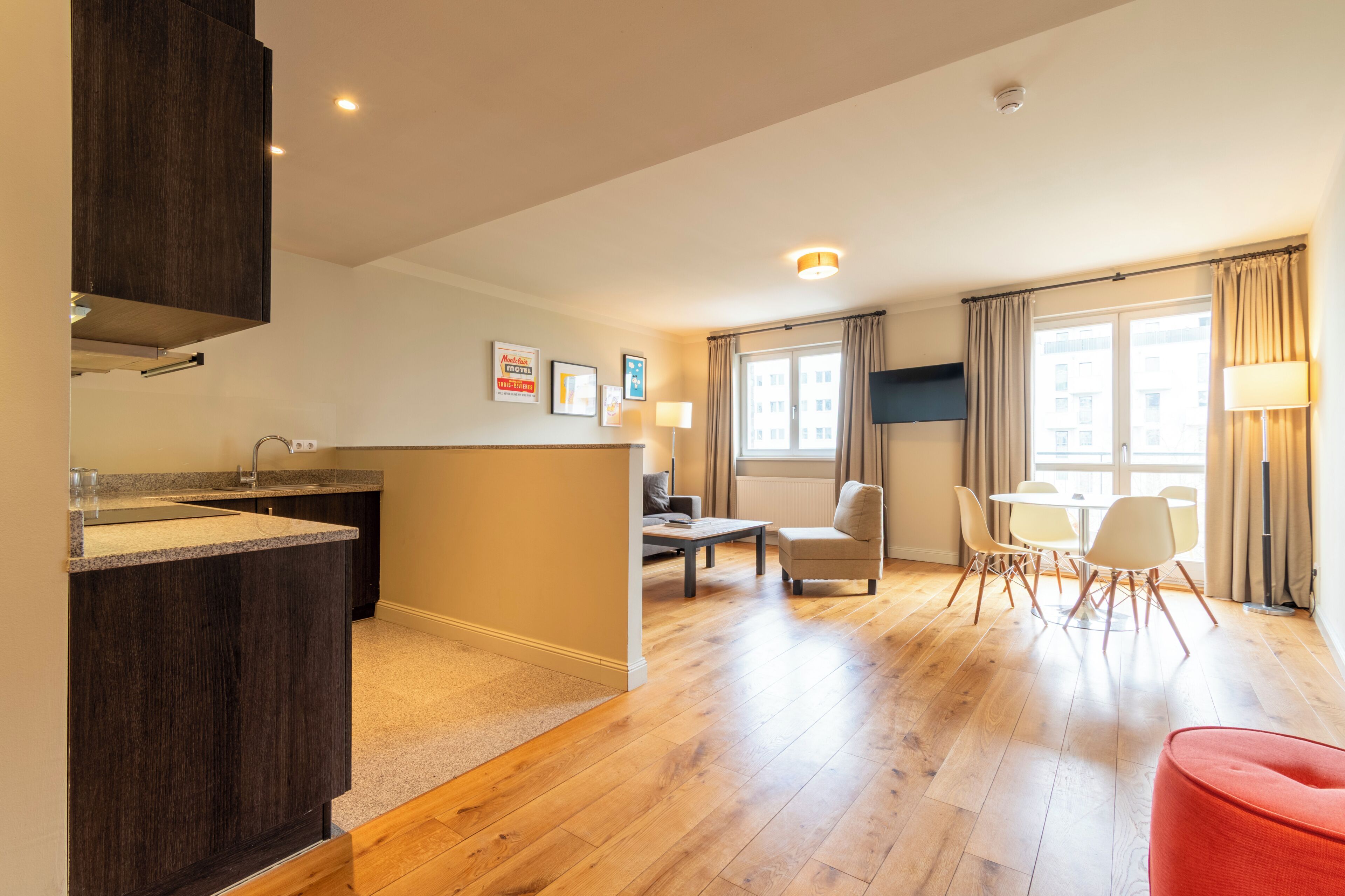 A modern kitchen with dark cabinetry, a dining area with a table and chairs, and a living space with a sofa and a red ottoman.