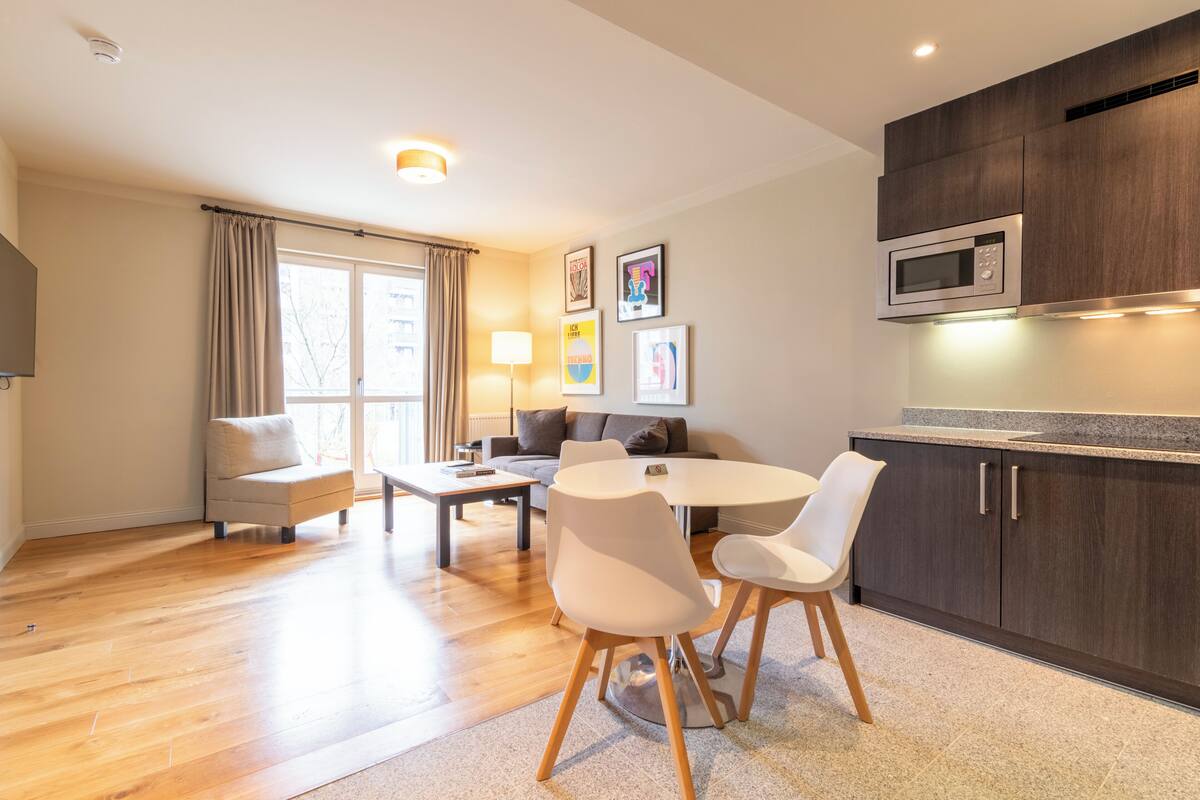 A modern living room with a dining area, a kitchen with dark cabinets and a microwave, and a large window with curtains.