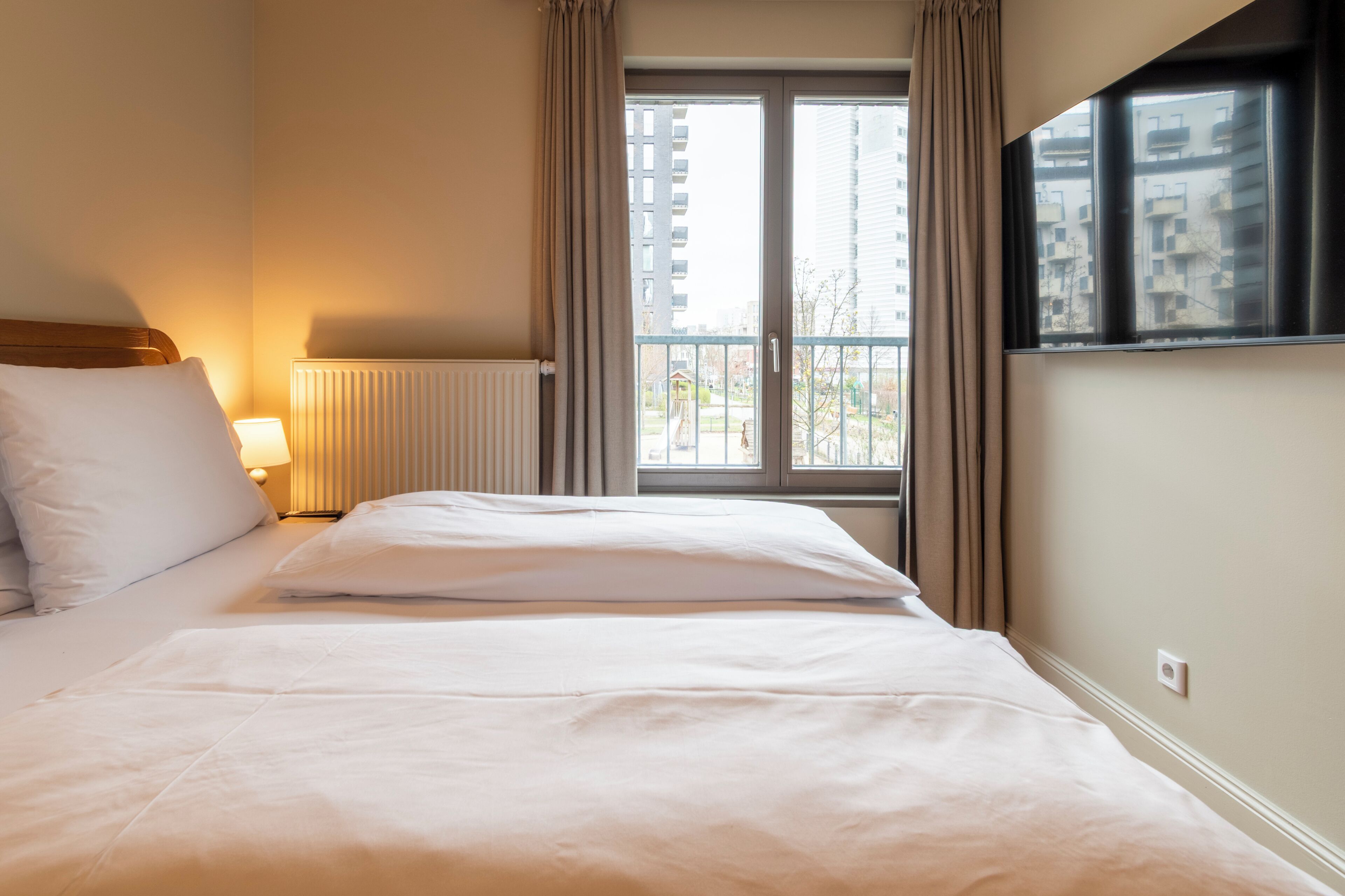 Habitación de hotel con cama, ventana con cortinas, radiador y vista a edificios.
