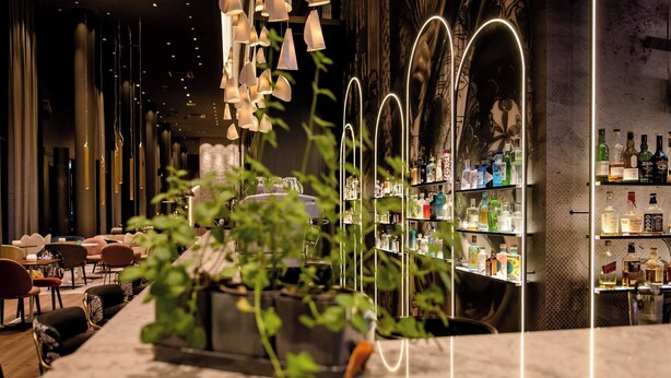 A bar area with a long counter, shelves of liquor bottles, hanging light fixtures, and potted plants. - Motel One Dresden Am Zwinger (Dresden)
