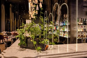 A bar area with a long counter, shelves of liquor bottles, hanging light fixtures, and potted plants. - Motel One Dresden Am Zwinger (Dresden)