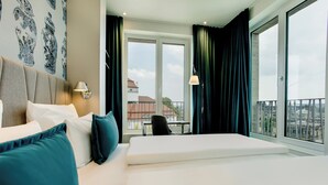 A hotel room with a large bed, a desk, a chair, and a view of the city through the windows. - Motel One Dresden Am Zwinger (Dresden)